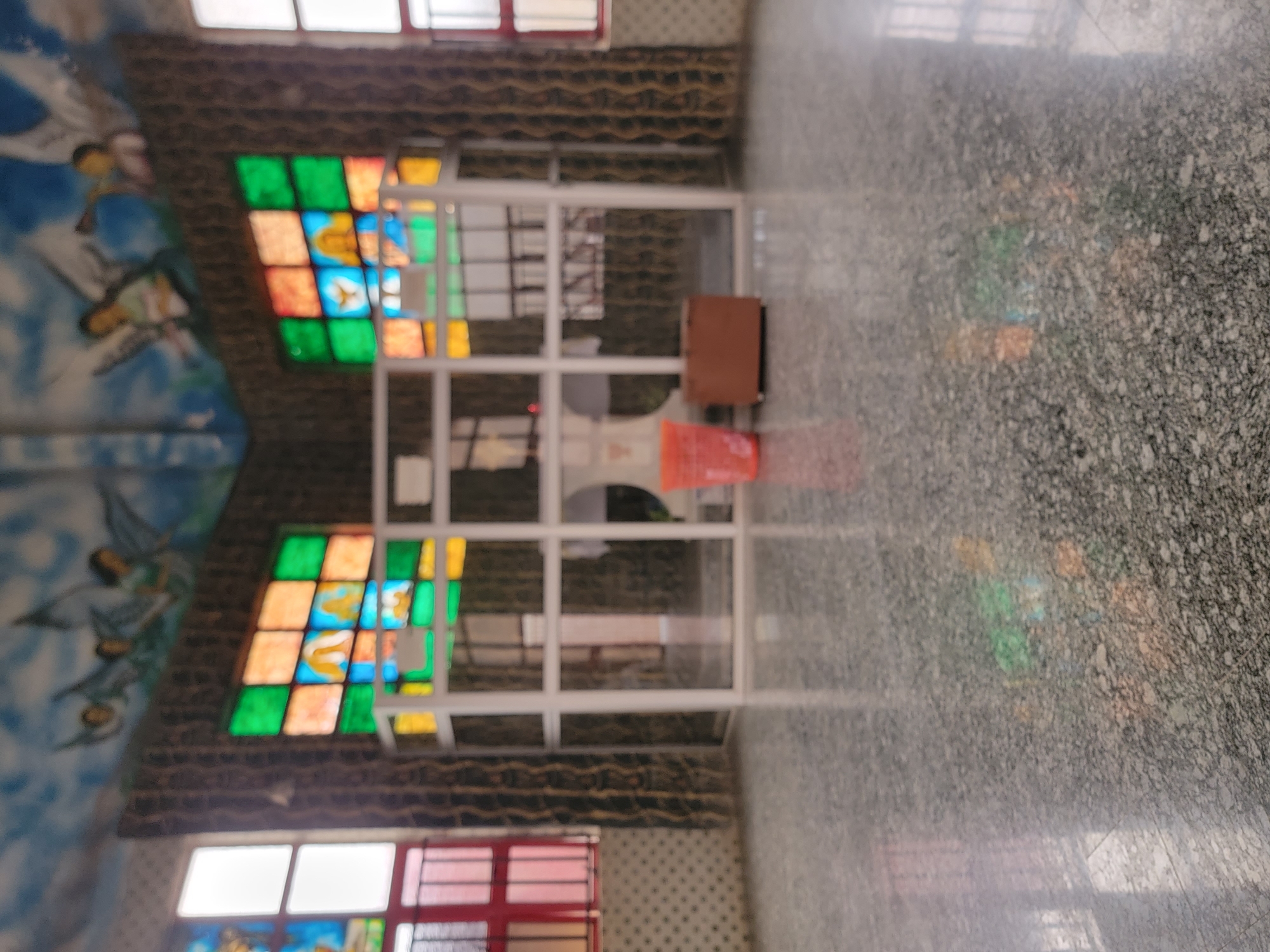 The image shows the interior of a building in Maiburiji, Nigeria. A white-framed window with colorful stained-glass panes is visible. The walls are decorated with a mural featuring figures, possibly angels, painted on a blue background. A brown cabinet and a red plastic container are positioned in front of the window. The floor is covered with a speckled gray material, possibly tile or concrete. The lighting is diffused. The scene is somewhat blurry.