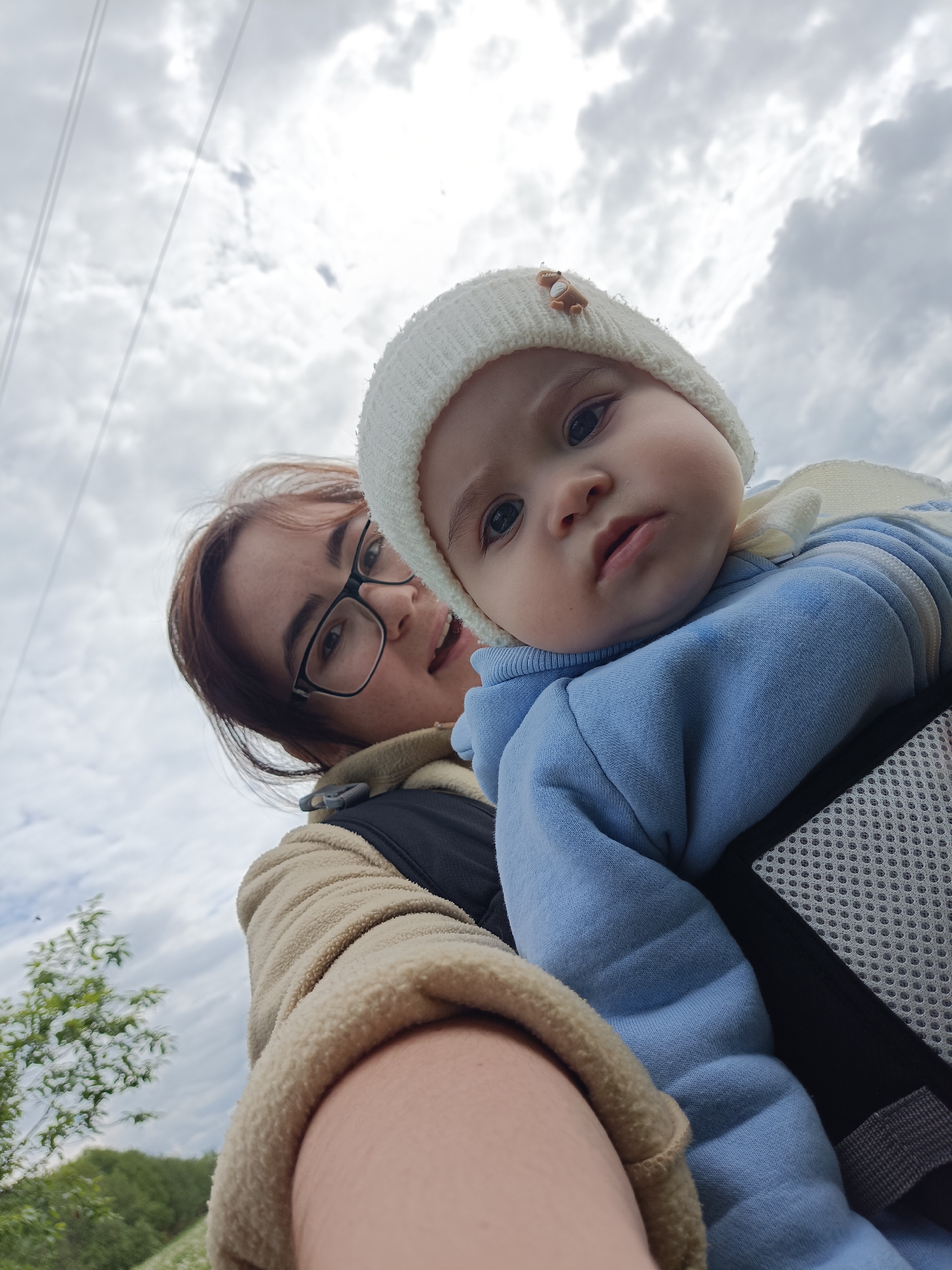 A selfie captured from a low angle shows a woman holding a baby in a carrier. The baby, wearing a light blue outfit and a white knitted hat with a small bear-shaped adornment, is the primary focus. The woman, with glasses and medium-length brown hair, is partially visible behind the baby. The background reveals a cloudy sky, with power lines visible in the upper left corner. A tree branch and hints of green vegetation can be seen in the lower left corner, suggesting an outdoor setting. The location is likely near Jelpachiha, Russia, given the presence of power lines and sparse vegetation. The woman appears to be holding the baby close, possibly on a walk or outing.