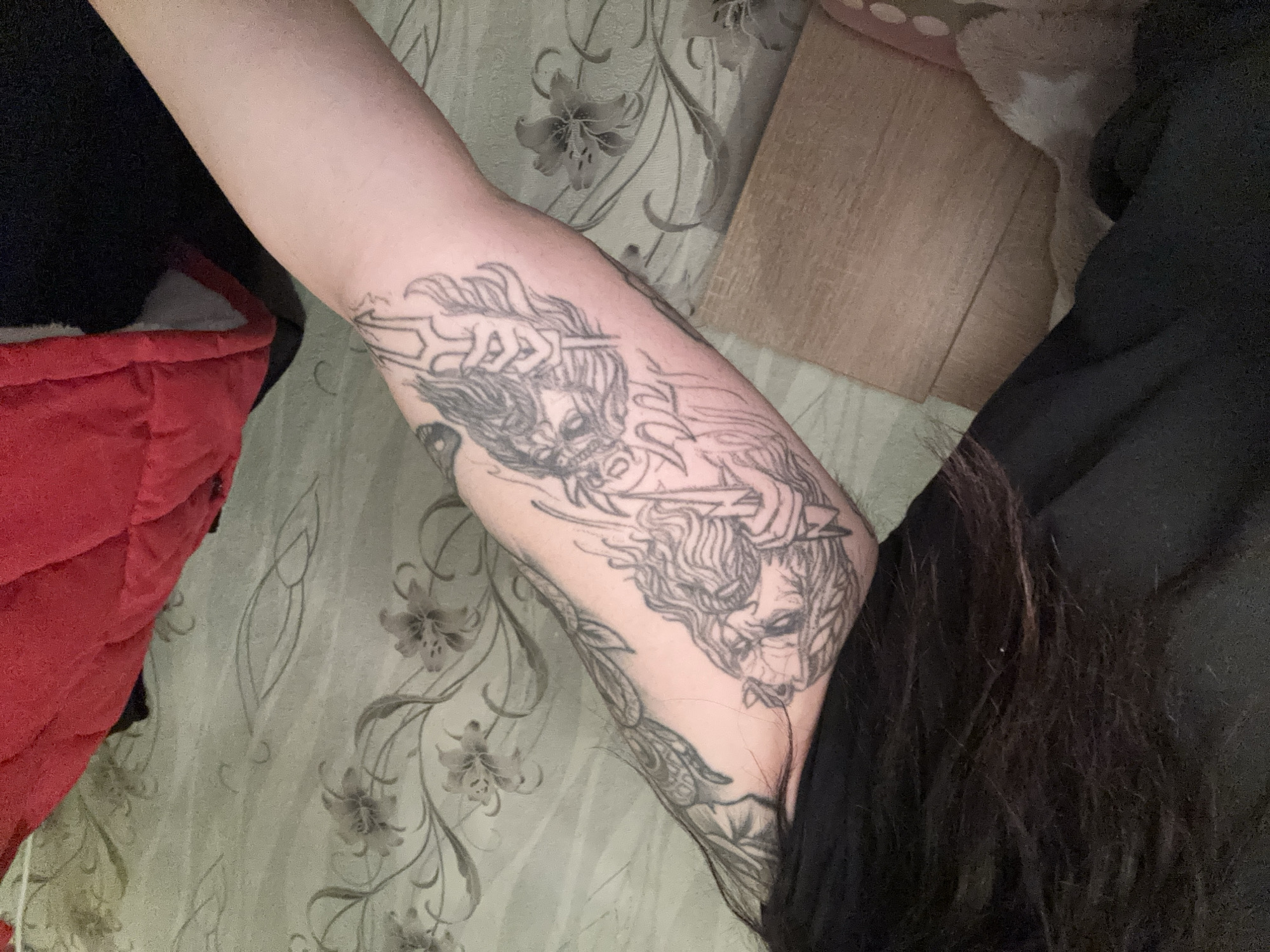 This scene takes place in an indoor setting within Dobrovnitsa, Bulgaria. The primary subject is a person's left arm, prominently displayed and extending from the upper left toward the lower right of the frame. The arm features a large, detailed, uncolored line-art tattoo on its outer forearm. The tattoo depicts an older, bearded male figure with long hair, holding a lightning bolt in his left hand. Below this figure, a second figure with headwear, possibly a crown or helmet, is depicted holding a trident or similar three-pronged weapon. Additional abstract or organic-patterned tattoos are visible on the person's upper arm, extending onto the shoulder area. The person's long, dark hair is visible on the far left, partially obscuring a black garment worn on the upper body. To the right, a segment of a red quilted jacket or vest is present. The background consists of a wall covered in light grey-green wallpaper adorned with a repeating pattern of dark outlined lilies and intertwining foliage. In the lower left corner, a soft, light-colored textile object rests next to a light brown, rectangular surface, possibly wood or laminate.