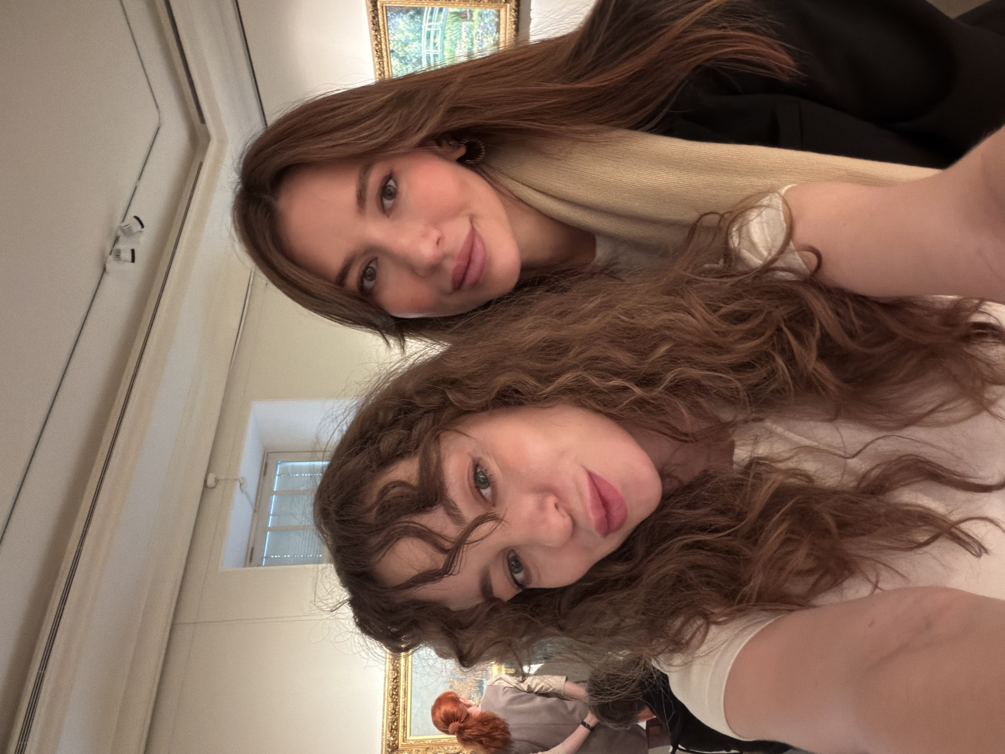 Two young women are taking a selfie in what appears to be an art gallery. The woman on the top has long, straight brown hair and is wearing a beige scarf and a black jacket. The woman on the bottom has long, curly brown hair and is wearing a white shirt. In the background, paintings hang on the wall, and there is a window. There are other people in the background as well. While a precise location in Moscow, Russia, cannot be verified from this image alone, the presence of art and the style of architecture suggest an art gallery setting in that city, or a similar location.