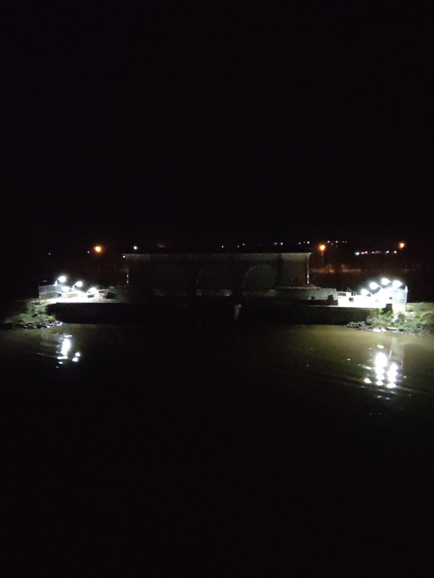 The image depicts a nighttime scene of a river in Yakhroma, Russia, with a structure spanning across the water. The structure appears to be an old bridge. The scene is illuminated by artificial lighting. Lights on the structure and along the distant shoreline cast reflections on the surface of the water. The sky is dark and there are no visible people or animals in the immediate frame. The image is dark, with contrast provided by the scattered lights.