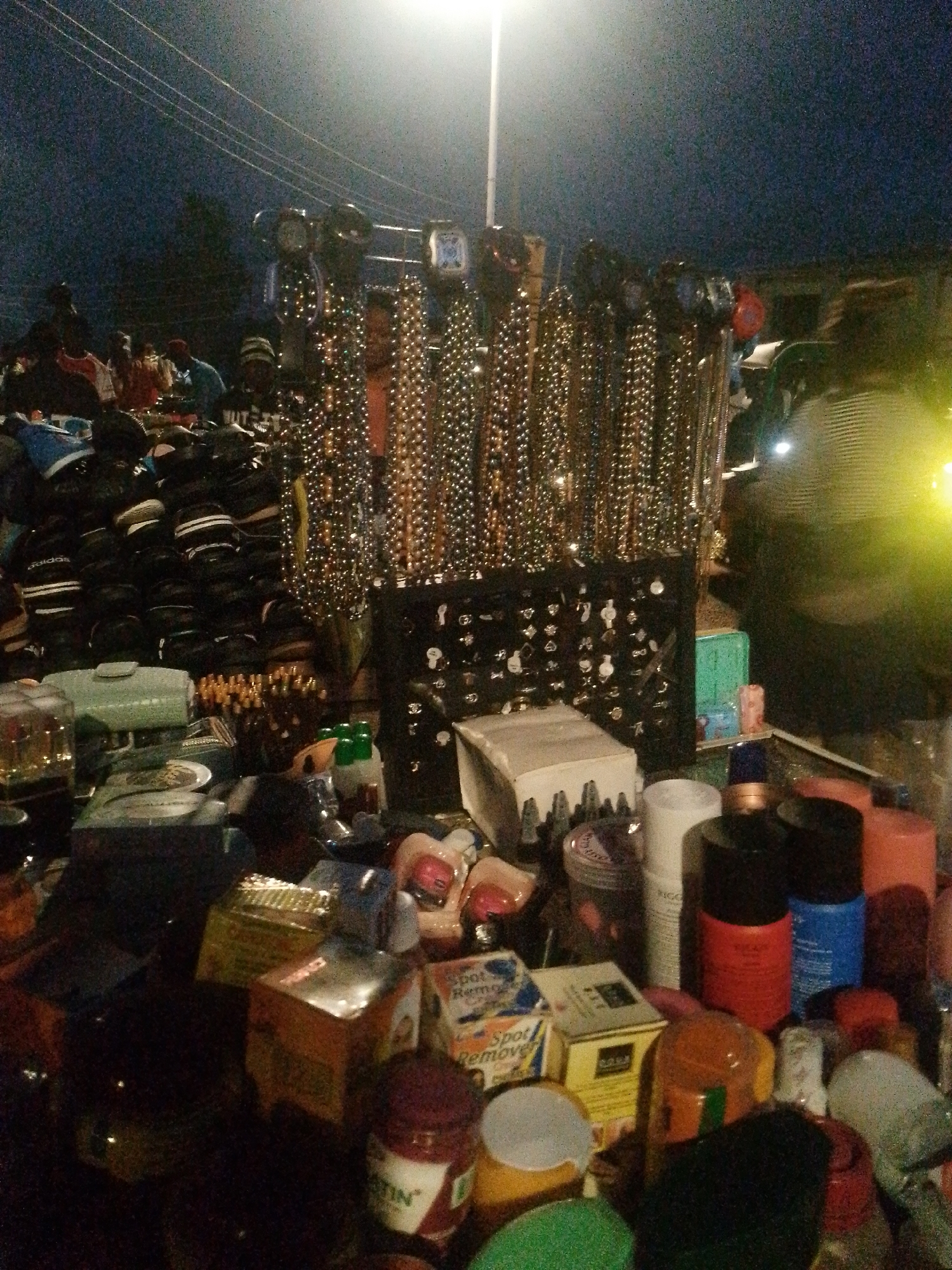 This is a medium-shot, vertical, night-time photograph of an outdoor market stall. The stall is displaying an assortment of goods, including necklaces, hats, and various toiletries or cosmetics. In the background, there are indistinct figures of people and overhead power lines, suggesting an urban setting. The lighting is artificial, with a bright overhead lamp and localized lighting sources that create highlights and shadows. The overall atmosphere is that of a bustling night market.