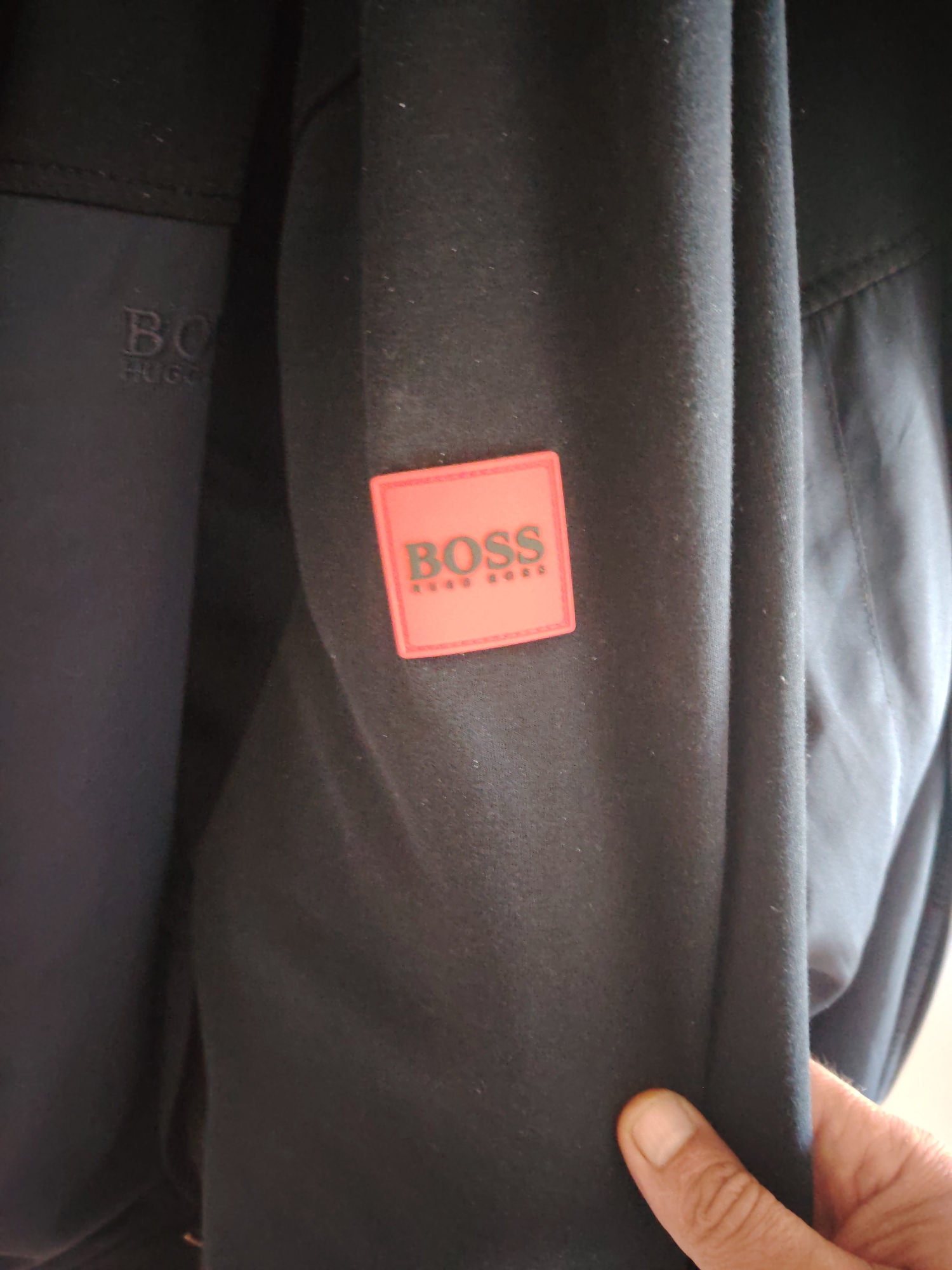 The image displays a close-up view of a dark-colored garment, likely a jacket or sweatshirt, worn by an unseen individual. On the right sleeve, a prominent rectangular patch, colored orange-red with a lighter border, features the brand name "BOSS" in dark, capitalized sans-serif letters, with "HUGO BOSS" written in smaller, stacked dark letters beneath it. To the left, on a darker, possibly contrasting fabric panel, a subtle, embossed or embroidered logo is partially visible, showing "BO" and "HUGO" in dark letters, indicating the "BOSS HUGO" brand. The fabric of the garment is a uniform dark tone, appearing soft and slightly textured. A human hand, with visible fingernails and thumb, holds the lower right portion of the garment, pulling it slightly taut. No other contextual elements, background, or specific location details are discernible within the frame.