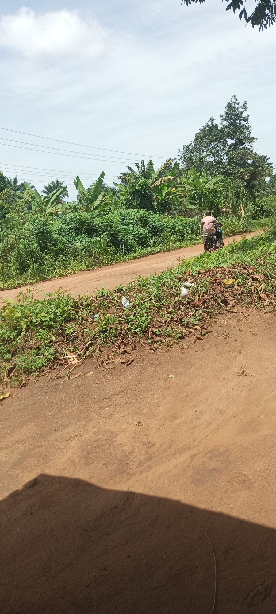 The image depicts a dirt road in a rural setting in Amauzu, Nigeria. A man rides a motorbike on the road. Lush green vegetation, including banana trees, lines both sides of the road. Scattered plastic debris is visible on the side of the road. Utility lines are strung across the sky. The foreground is a dirt area with some sparse vegetation and the shadow of the camera.