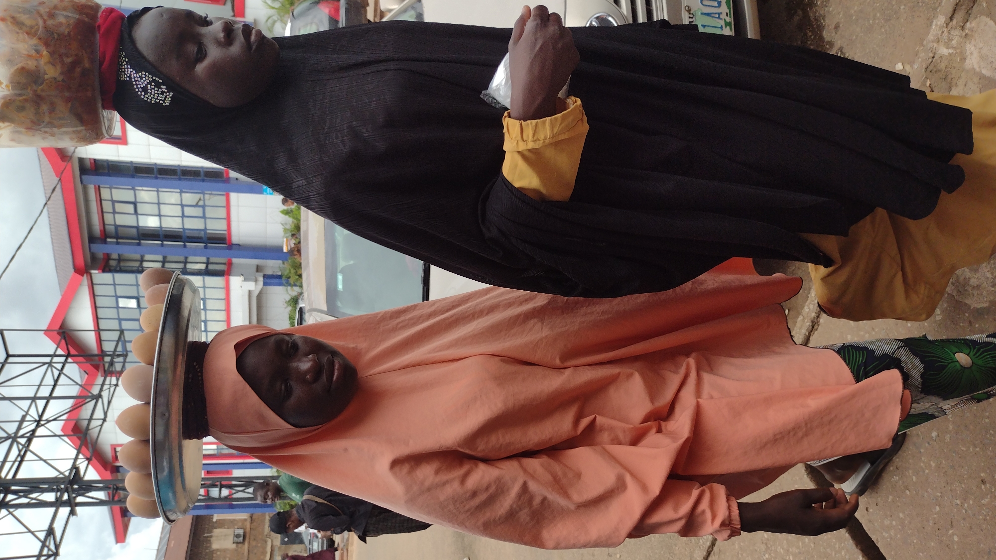 Two young women, likely vendors, are the subjects of this vertical, eye-level shot. The woman in the foreground wears an orange headscarf and a peach-colored dress. She carries a metal tray on her head, laden with at least ten eggs. Her face is turned towards the camera, and she appears to be looking slightly up and to her right.
Behind her, the second woman is dressed in a black headscarf and a black abaya with yellow cuffs. She wears a red headscarf beneath the black one, and a jar of food is perched on top of her head. Her hands are clasped together, holding what appears to be a plastic bag. She is looking away from the camera.
The background features a building with blue and red accents, suggesting an urban environment. A white car with a license plate is partially visible. The ground appears to be a dusty road. The lighting suggests it is daytime, possibly overcast, as there are no strong shadows. The overall impression is that of a busy street scene in Jos, Nigeria, where these women are likely engaged in selling goods.