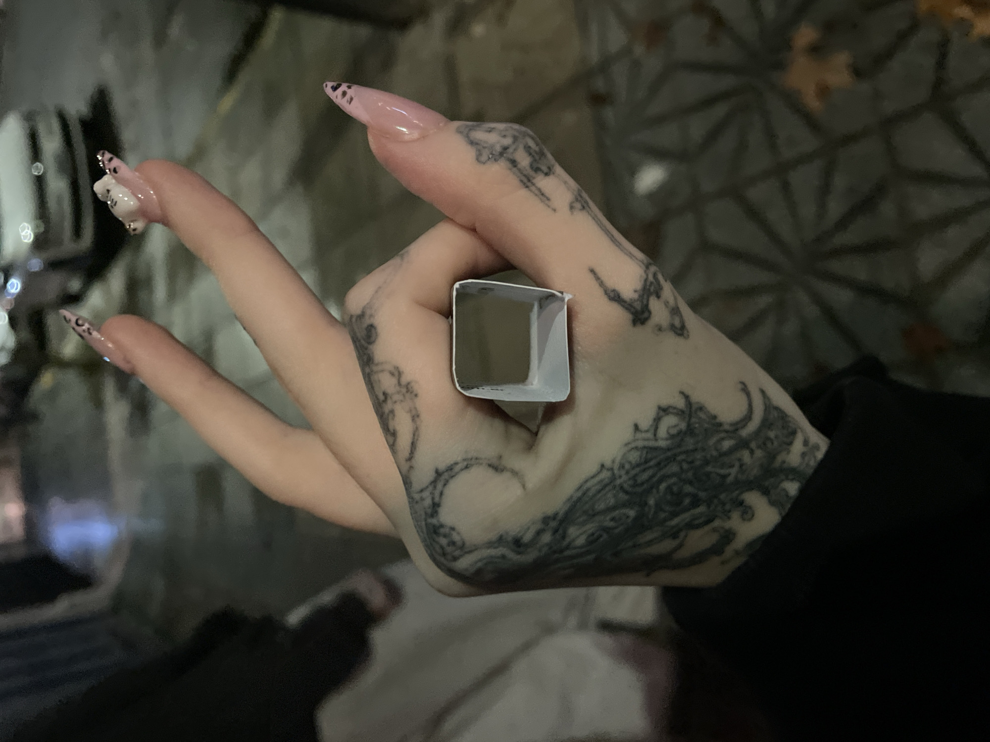A tattooed hand holds a small, square, white object. The hand's nails are long, pointed, and painted pink. The background appears to be a street in Buenos Aires, Argentina, with patterned stone pavement. A blurred light source is visible in the upper left corner. The scene suggests an urban environment, possibly at night.