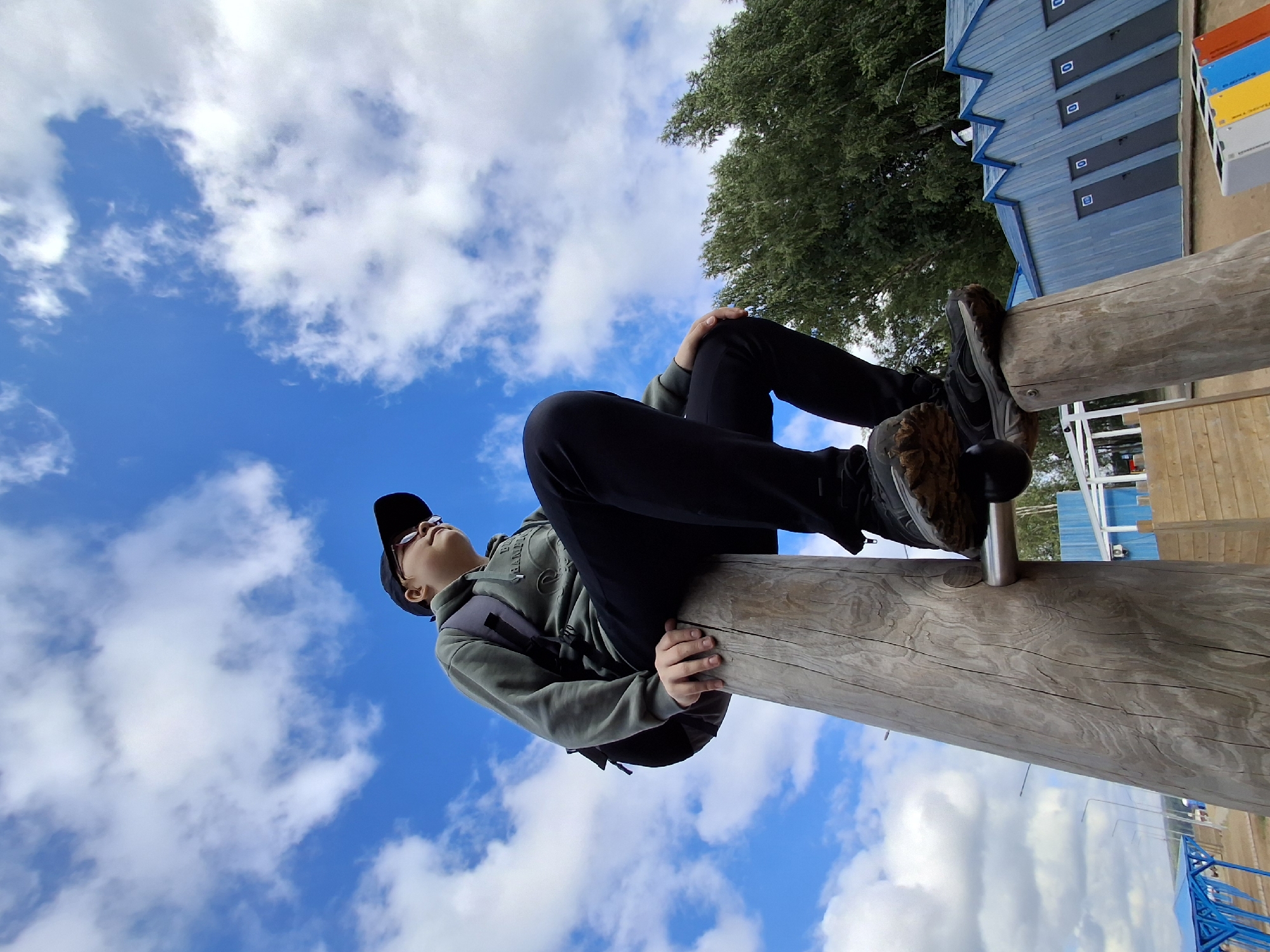 In Chaikovsky, Russia, a young person wearing a green hooded sweatshirt, black pants, a black baseball cap, and sunglasses is sitting on top of a large wooden pole. The person is looking up and to the right, with their hands resting on their knees. They are wearing a dark backpack. The wooden pole is part of a playground structure. In the background, there are trees, a blue building with multiple doors labeled with numbers, and a bright blue sky with white clouds. The ground appears to be sandy or dirt.