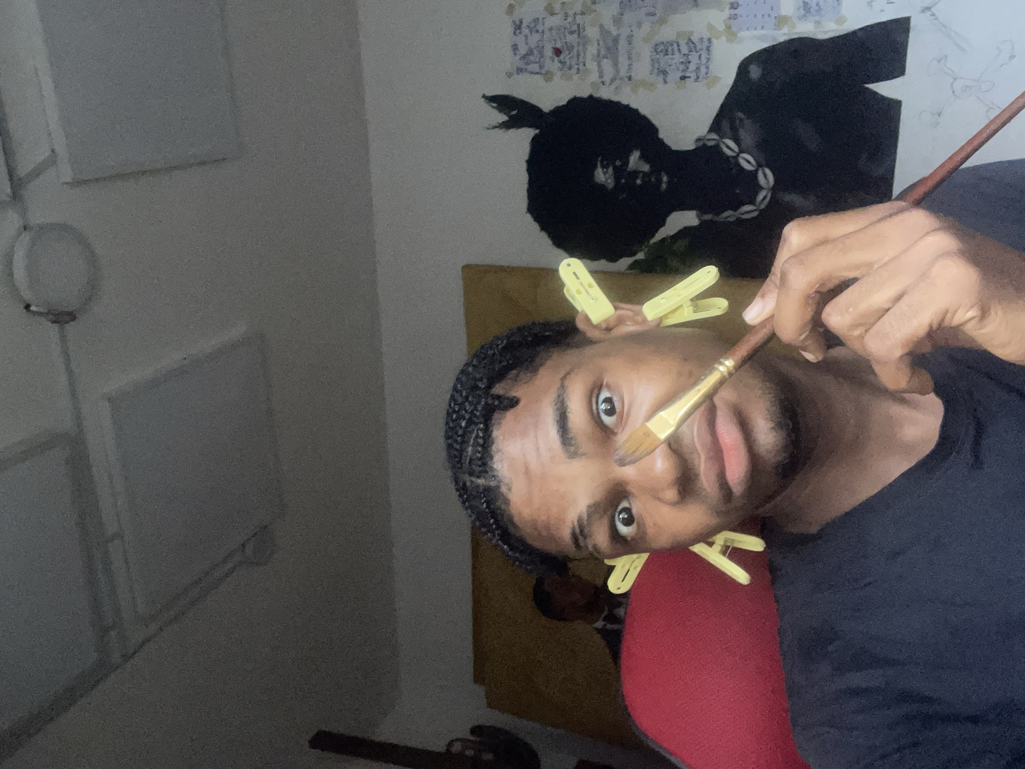 The image shows a young Black man with braided hair, holding a paintbrush to his nose and with yellow clothespins clipped to his ears. He is wearing a black t-shirt and is seated in front of a red chair. In the background, there is a silhouette of a person with an afro and a seashell necklace, as well as a painting or poster on the wall. Another person is visible in the background, wearing a suit. The environment appears to be an indoor space, possibly an art studio, with fluorescent lighting panels visible on the ceiling. There is no indication of the location being in Ado, Nigeria.
