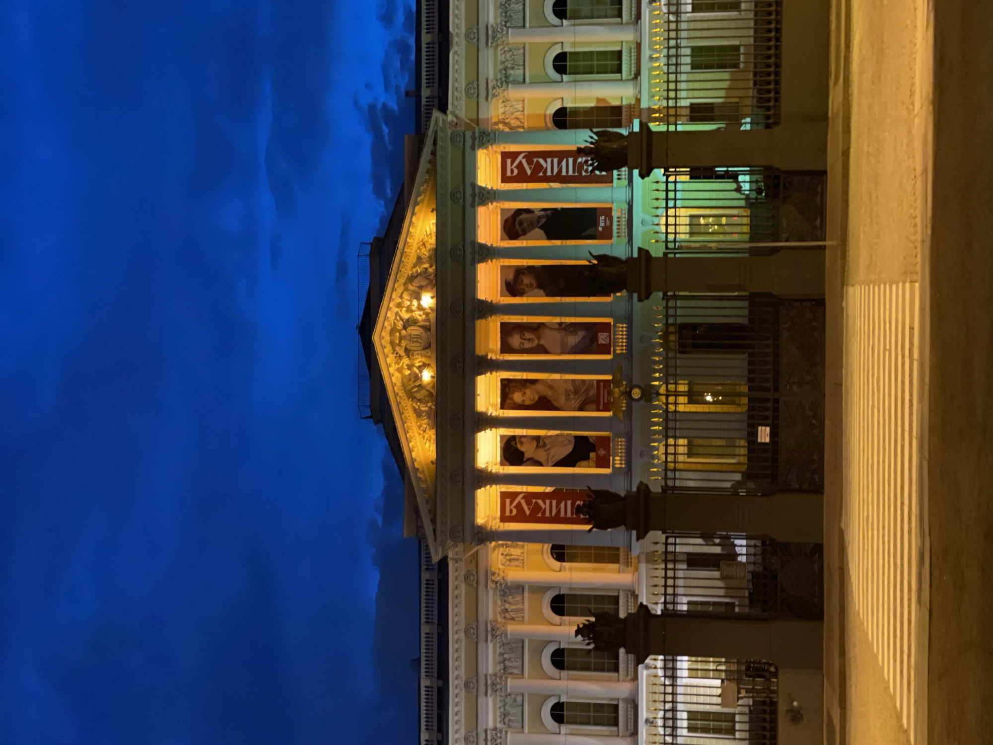 The image displays a majestic, classical-style building facade illuminated at night or during late twilight in Saint Petersburg, Russia. The grand architecture features numerous ornate columns, elaborate friezes, and a detailed pediment adorned with sculptures, all bathed in a warm yellow light that sharply contrasts with the deep blue, cloudy sky above.
On one of the upper levels, large red banners are prominently displayed within the arched openings between columns. These banners feature individual portraits, likely advertising an exhibition, performance, or cultural event. The Cyrillic text "КУЛИГА" (KULIGA) is clearly visible on the red banners. Smaller logos, including "ВТБ" (VTB), are also discernible on some of the portrait posters.
In front of the building, a dark metal fence or gate with decorative elements runs along the facade, partially obscuring the lower windows. To the right, a light-colored paved area or sidewalk is visible, showing distinct vertical lines from the artificial illumination. The dramatic lighting and imposing architecture create an atmosphere of cultural significance and historical grandeur, characteristic of Saint Petersburg's iconic buildings.