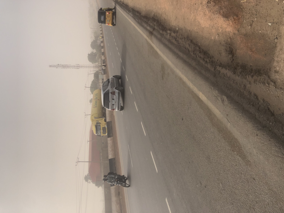 An asphalt road curves slightly with a white dividing line. A motorcycle with two people is in the foreground, followed by a silver car and a yellow truck with “Green Ville Lux” on it. Further ahead is a yellow tricycle. A cell phone tower rises beside the truck and car. Hazy or foggy conditions obscure the distant background. The shoulder of the road is dirt and gravel.