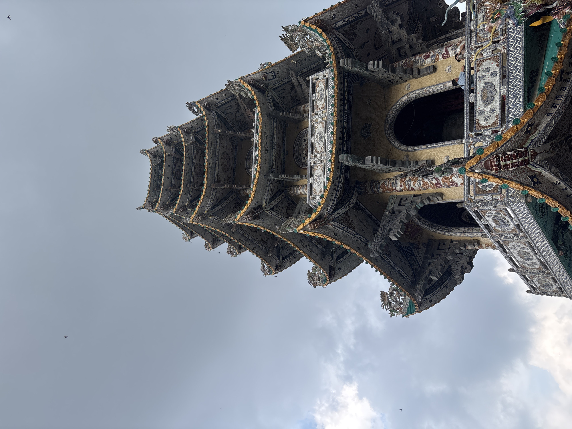 The image, rotated 90 degrees clockwise, displays a tall, multi-tiered, and highly ornate building, likely a pagoda or temple tower, against an overcast sky. The structure is richly decorated with intricate mosaic-like patterns, possibly crafted from ceramic or glass shards, covering its walls and tiered rooflines. Golden accents highlight the edges of each tier and various architectural details. Sculptures resembling dragons or mythical creatures adorn the corners of the eaves.
Through one of the building's arched openings on an upper level, a person's head and shoulders are partially visible, looking outwards. The sky is largely overcast with light gray clouds, indicating daytime. Several small, dark birds, resembling swallows, are seen flying in the sky, particularly in the upper left quadrant. No clearly legible text is identifiable, though some decorative patterns might incorporate stylized characters. The architectural style is consistent with traditional temple structures found in Xuân Thành, Vietnam. The perspective looks upwards, emphasizing the grandeur and intricate craftsmanship of the building.