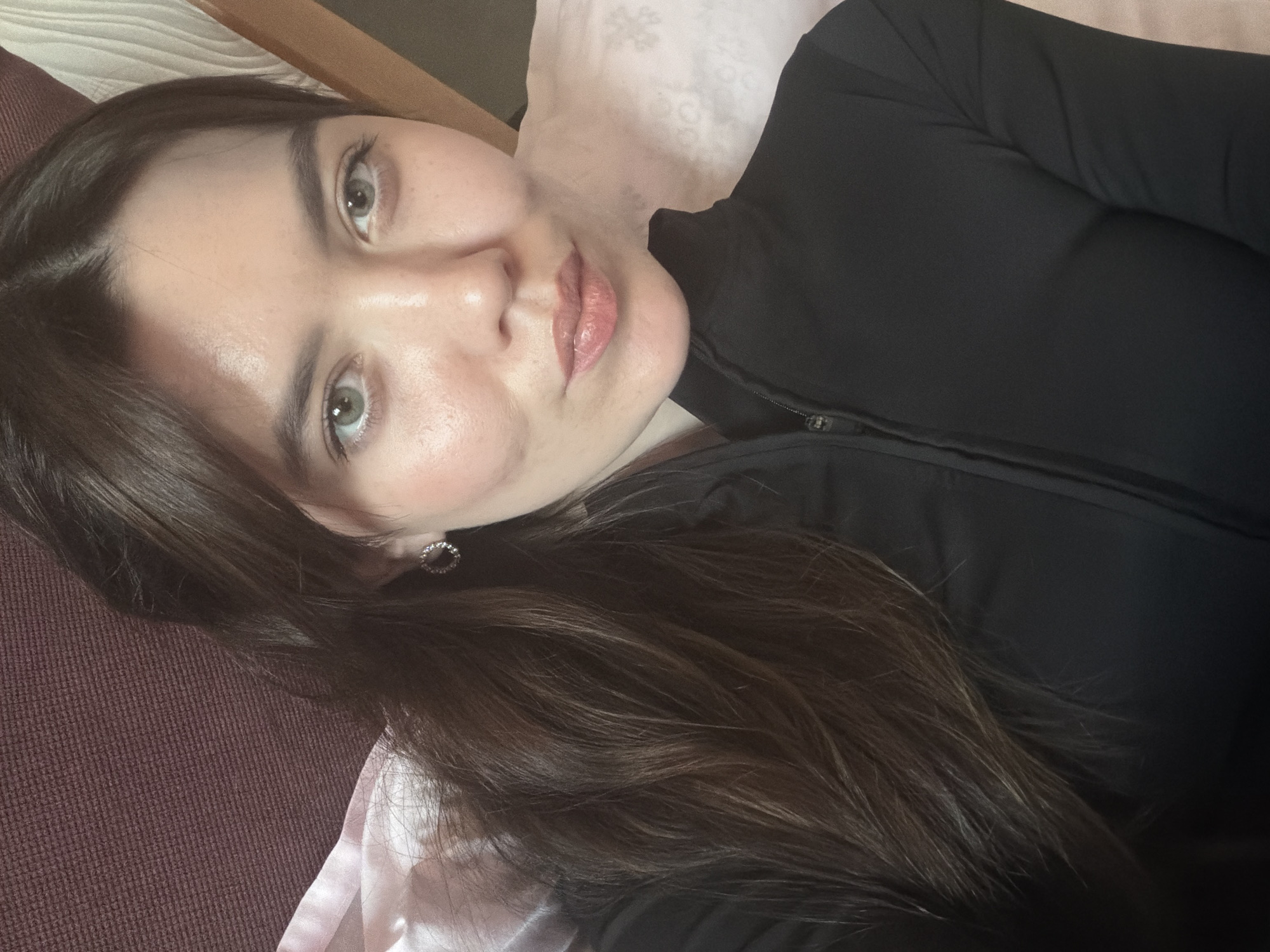 A selfie of a woman with long brown hair and green eyes is captured in an indoor setting. She wears a black zip-up garment. An earring is visible on her left ear. She is lying on what appears to be bedding, including a brown headrest and a pink pillow. The location is not discernible from the image.