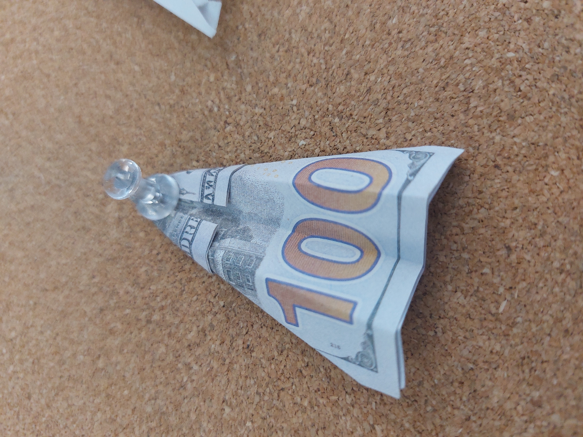 The image shows a paper airplane made from a U.S. one hundred dollar bill, pinned to a brown corkboard with two clear pushpins. The location is not apparent in the image, and it is therefore not possible to determine if the image was taken in Burgas, Bulgaria.