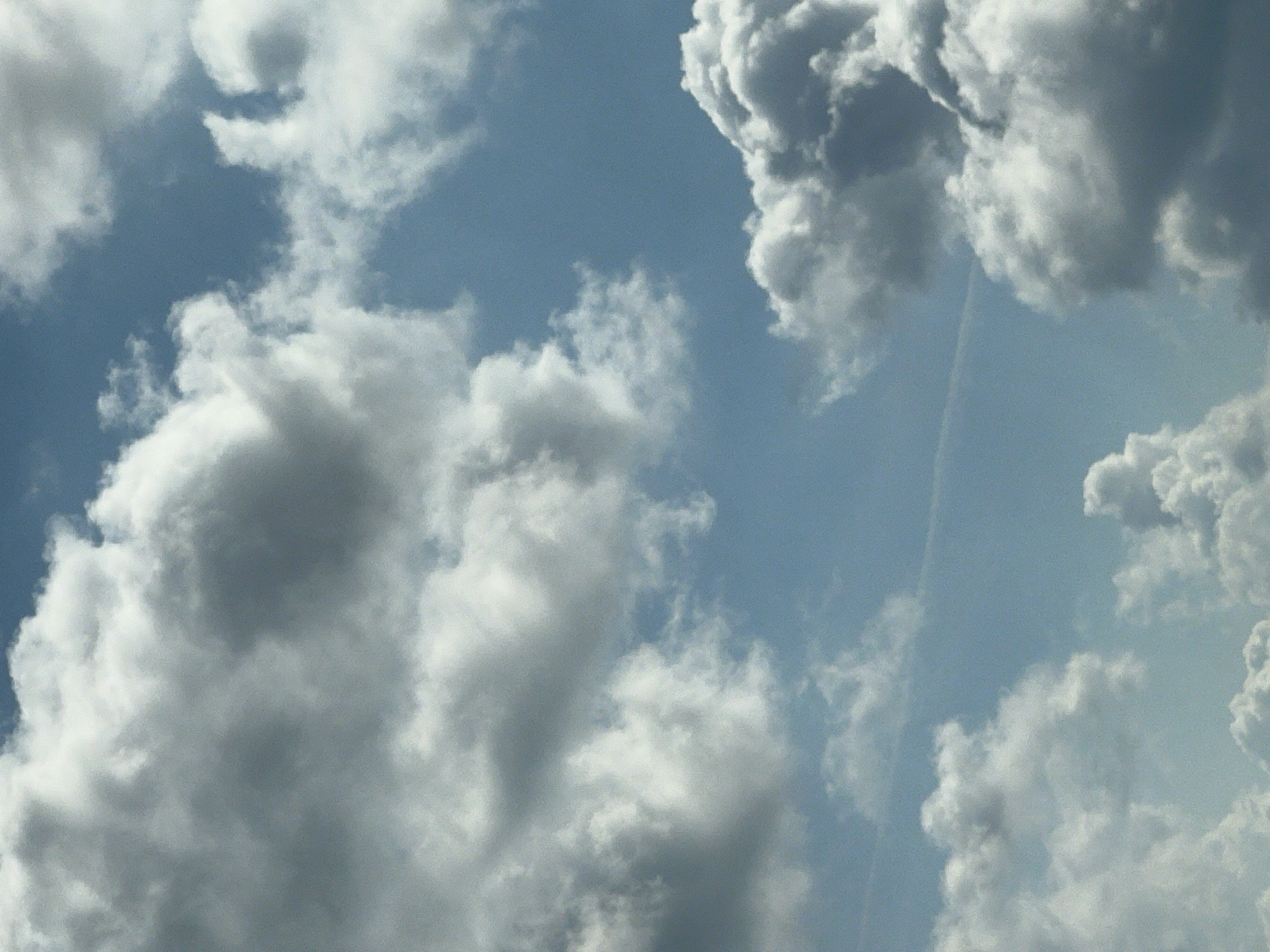 The image shows a view of the sky filled with fluffy, white and gray clouds against a backdrop of clear blue sky. There is a faint contrail visible in the upper left quadrant of the image, and another thinner contrail near the center of the image stretching from left to right. The lighting suggests it is daytime, possibly late morning or afternoon, with the sun illuminating the clouds from above and to the right, creating shadows and highlights that give them depth and texture. No people, buildings, or other recognizable structures are visible. The image is an outdoor, natural scene, devoid of urban elements, making it impossible to determine a specific location within Warsaw, Poland, or any other city.