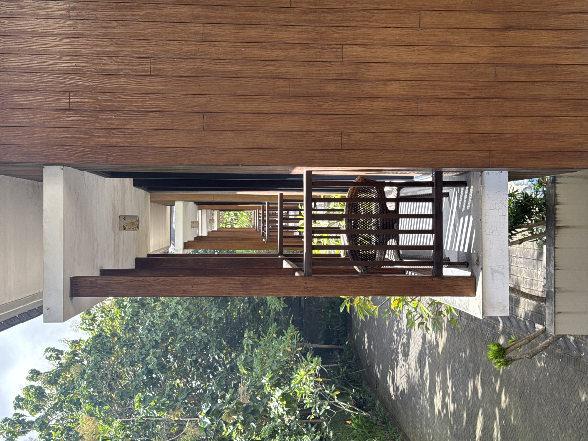 The image depicts a long, narrow perspective of an outdoor area in Kuta Selatan, Indonesia. A series of repeating balconies stretch into the distance, lined with wooden railings and furniture. One visible chair is of a wicker design. Above the balconies, there is a brown, wooden exterior wall, possibly of a building. Below, the scene is dominated by lush green trees and a paved area that creates dappled light and shadow patterns. The overall impression is one of a tropical setting.