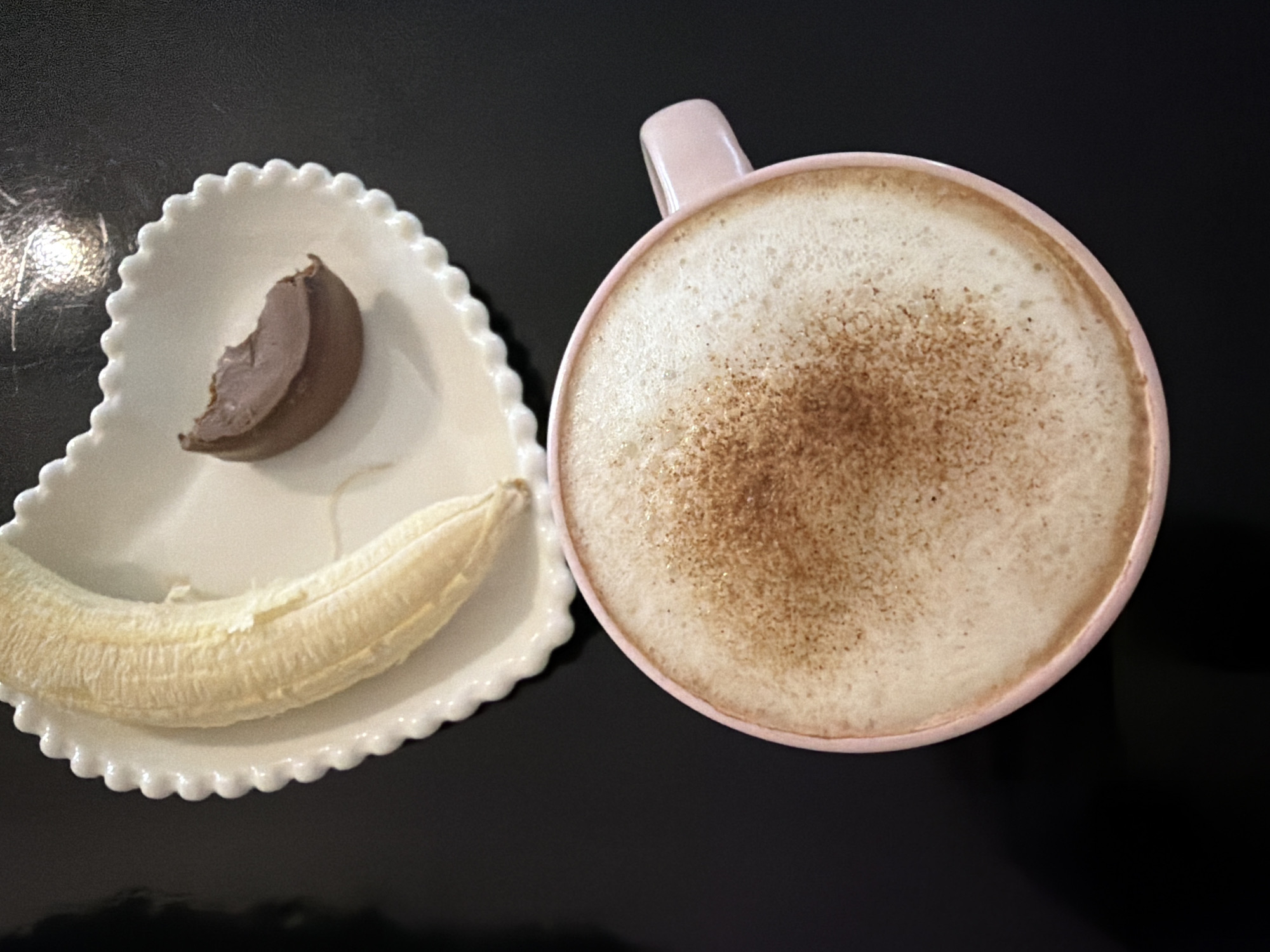 A top-down view shows a light pink mug filled with a foamy beverage sprinkled with a brown powder (likely cinnamon or cocoa) and a heart-shaped plate holding a peeled banana and a partially eaten chocolate piece. The items rest on a dark, reflective surface. The location of Pervomaysky, Russia, is not identifiable from the visual content.