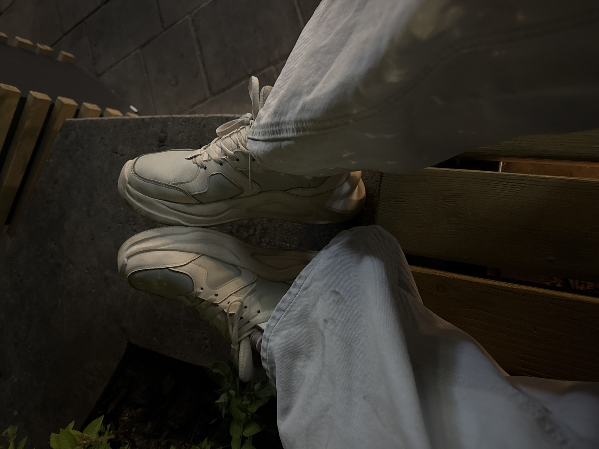 A perspective image shows a person's legs and feet, clad in loose white denim jeans and white sneakers, resting on an outdoor bench. The background suggests an urban environment, possibly in the city of Kazan, Russia. The sneakers are clean with a thick sole. The person is situated next to a concrete structure and wooden slats, with a hint of greenery in the lower left corner. The lighting is dim, indicating either evening or a shaded area.