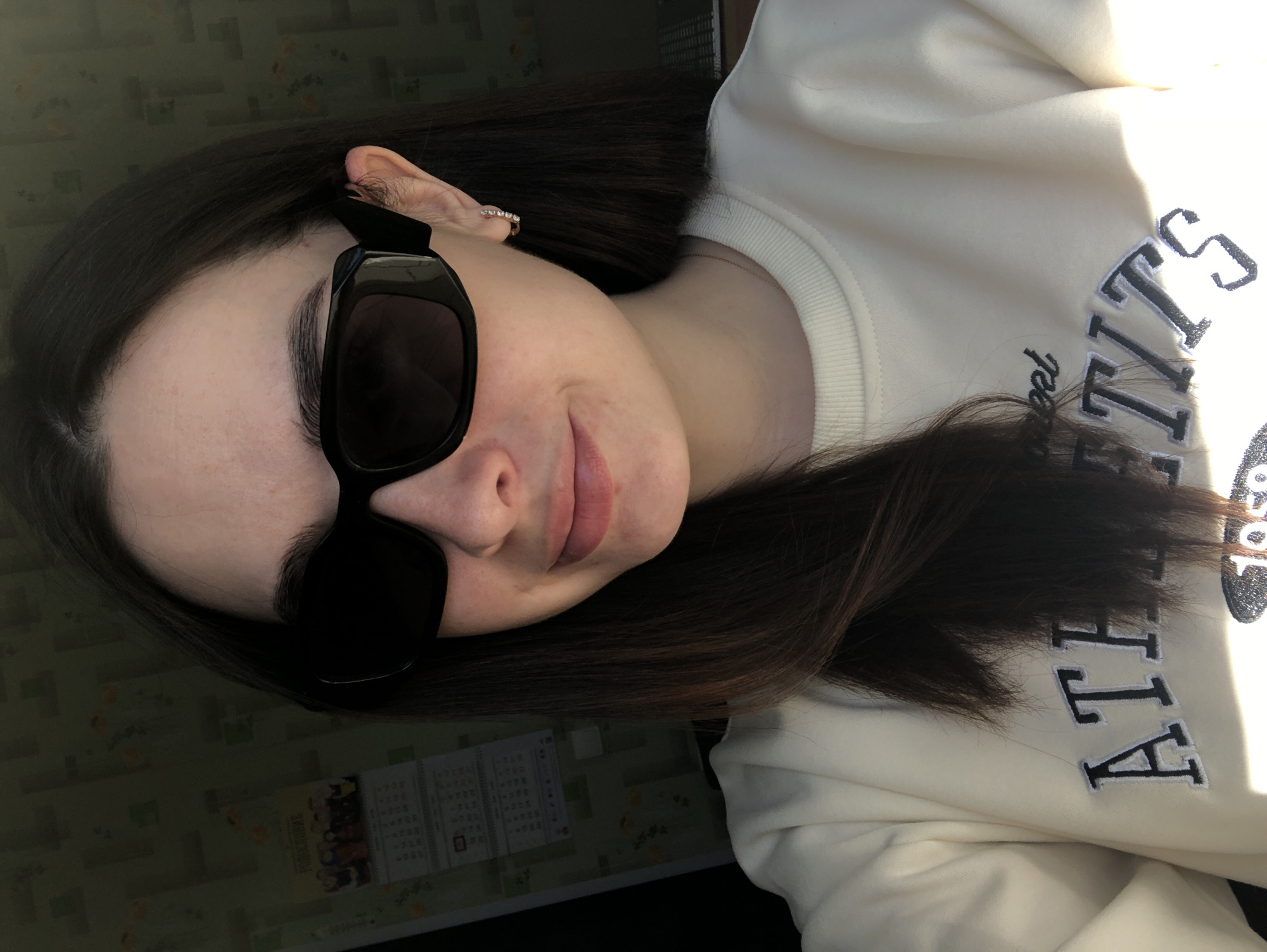 A woman with long, dark hair is wearing black sunglasses and a light-colored sweatshirt. The sweatshirt has the word "ATHLETICS" printed on it. She is wearing an earring. A calendar or bulletin board is visible in the background. The location is not determinable from the image.