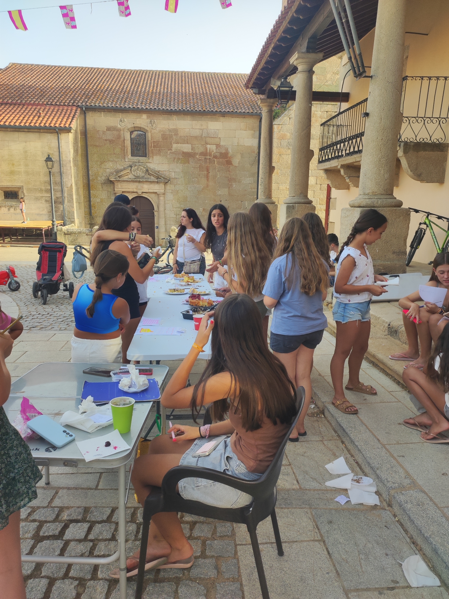 The image depicts an outdoor gathering of young people, primarily girls, in the town square of Sobradillo, Spain. A long, white table is set up, laden with food, including cakes and pastries, and drinks in cups. Several individuals are gathered around the table, some holding plates or engaging in conversation. In the background, the distinctive architecture of Sobradillo's town square is visible, with stone buildings, arched walkways supported by large columns, and a tiled roof. The ground is paved with cobblestones. A red and black stroller and a green bicycle are also present in the scene, suggesting a casual, community event. The atmosphere appears lively and social, with people interacting and enjoying the food and company.