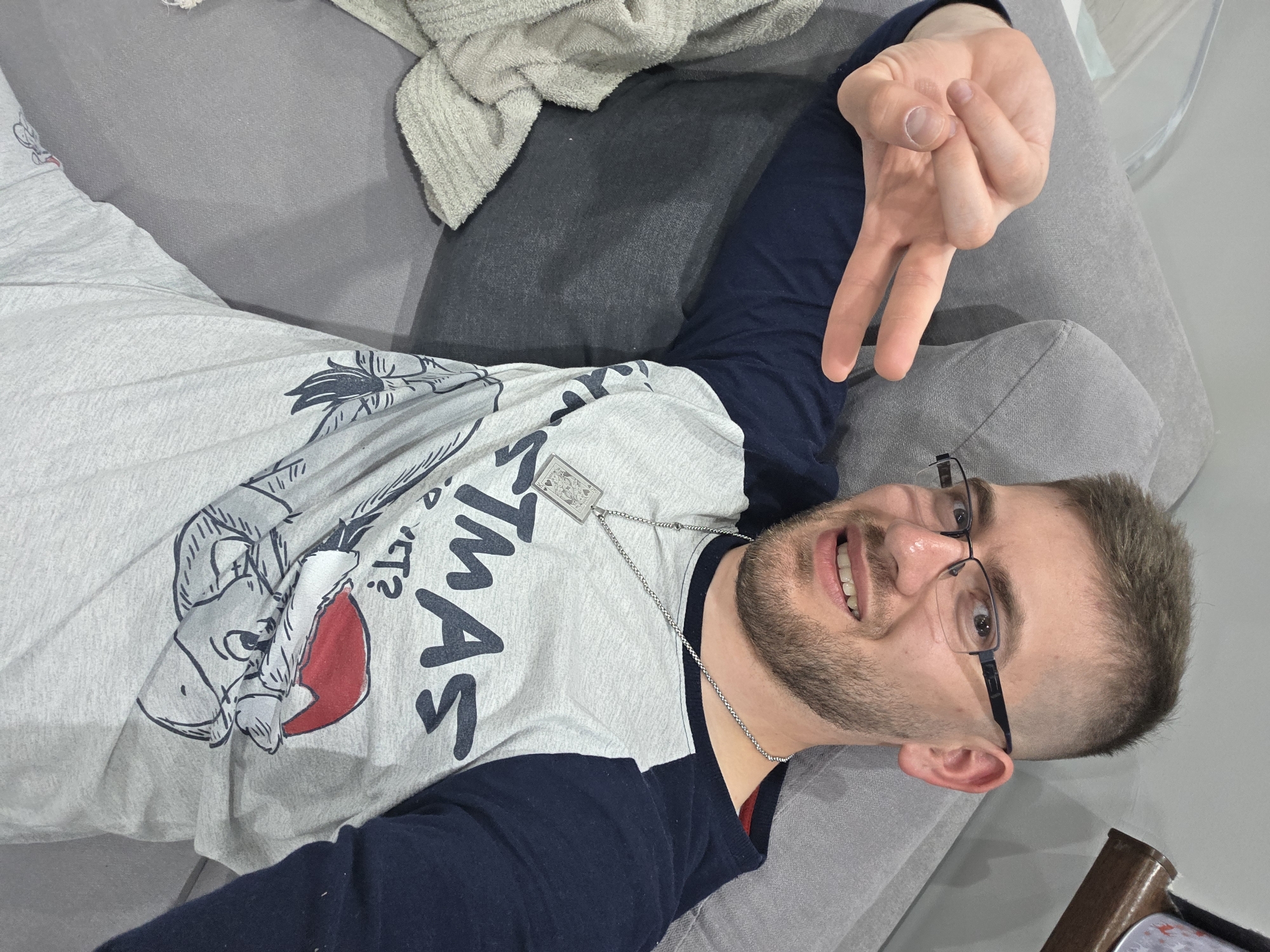 A man with short brown hair, a beard, and glasses is lying on a gray couch. He is wearing a gray and navy blue long-sleeved shirt with a graphic on the front, and a silver chain necklace with a pendant. He is holding up his hand in a peace sign. A folded cream-colored towel rests on the couch near his head. The background is a light gray surface. The location is not Yagodovo, Bulgaria.