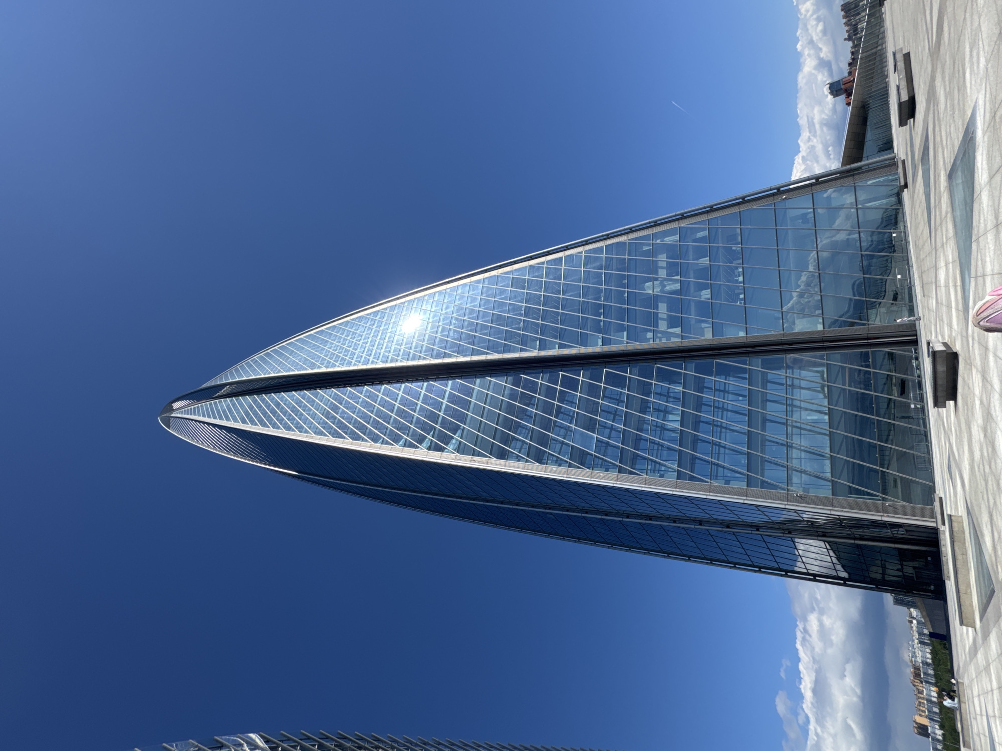 This image, originally oriented vertically, captures a striking modern architectural scene under a bright, clear blue sky. The central subject is a towering, slender skyscraper with a distinct curving or twisting form, predominantly clad in reflective glass panels arranged in a grid pattern. Intense sunlight creates a brilliant glare on the building's upper facade, reflecting the deep blue sky.
To the right of this prominent structure, a paved outdoor area, possibly a plaza or rooftop, is visible, alongside an adjacent building that also features reflective glass elements. This adjacent structure reflects distant white clouds and a low-rise city skyline on the horizon. A small portion of a person's pink and purple shoe is visible on the paved area in the top right corner, indicating a human presence and the vantage point from which the photograph was taken.
The scene conveys a sense of modernity and openness, set against a backdrop of a vast, unblemished blue sky with scattered clouds primarily concentrated near the horizon. The bright light suggests a daytime setting, likely midday or early afternoon, with excellent weather conditions. No specific activity beyond observation is occurring, and there is no visible text. The architecture is indicative of contemporary urban development, located in Saint Petersburg, Russia, as per the provided context.