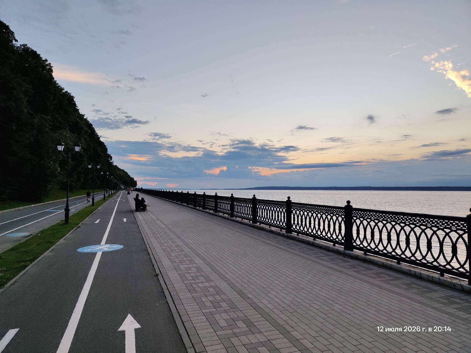 This is a wide, horizontal, eye-level, medium-shot photograph of a waterfront promenade at dusk. The sky is a pale blue with streaks of pink and orange from the setting sun, and a few scattered clouds. The promenade is paved with light grey bricks laid in a basketweave pattern. A black, ornate metal fence runs along the edge of the promenade, separating it from the water. The water is a calm, greyish-blue, reflecting the sky. On the left side of the promenade is a bike path, marked with white lines and a blue circular sign with a pedestrian symbol. A grassy embankment with trees rises sharply to the left of the bike path. Several lampposts line the embankment. In the middle ground, on the promenade, a couple is sitting on a bench. Further in the distance, a few other figures are visible walking. The bottom right corner of the image contains a timestamp: "12 июля 2026 г. в 20:14".