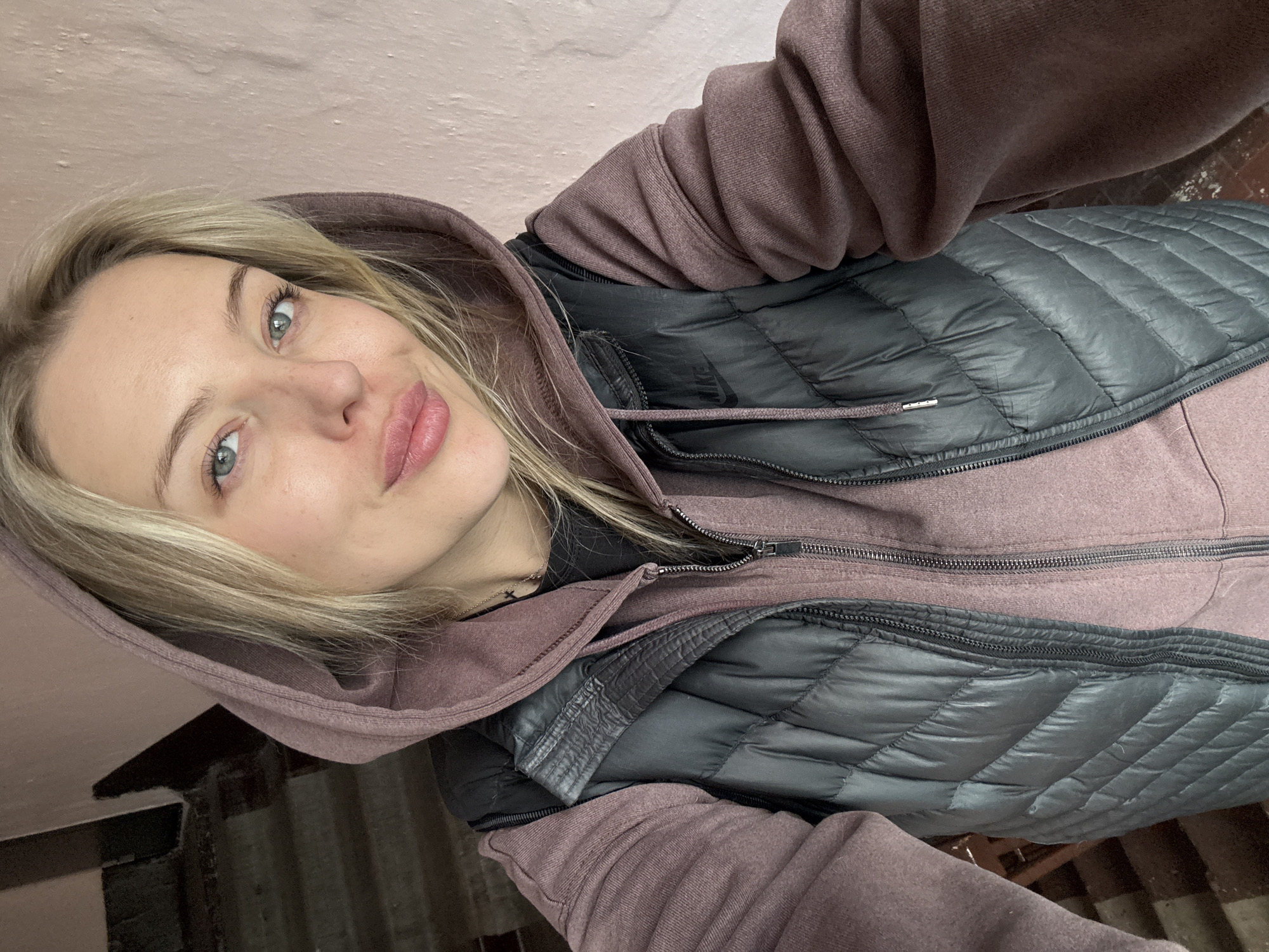 A young woman with blonde hair and light blue-green eyes is captured in a close-up, selfie-style photograph. She looks directly towards the camera with a slight, relaxed smile or smirk. Her lips appear full with a hint of pink color, and her eyelashes are defined with mascara. She is wearing a mauve or dusty pink hooded sweatshirt, which has its hood pulled up around her head, and a dark grey or black quilted vest layered over it. Both the sweatshirt and the vest have visible zippers. A thin silver chain with a small cross pendant is visible around her neck. On the dark vest, the brand name "NIKE" is discreetly visible.
The setting appears to be indoors, possibly within a building or a stairwell in Borzya, Russia. The background consists primarily of a textured, light pink or peach-colored wall behind her. To the lower left and right, darker, indistinct shapes suggest architectural elements such as stairs or corners, providing a utilitarian rather than decorative backdrop. The lighting is even, suggesting indoor conditions rather than specific outdoor time of day or weather. The overall mood conveyed by the subject is calm and confident.