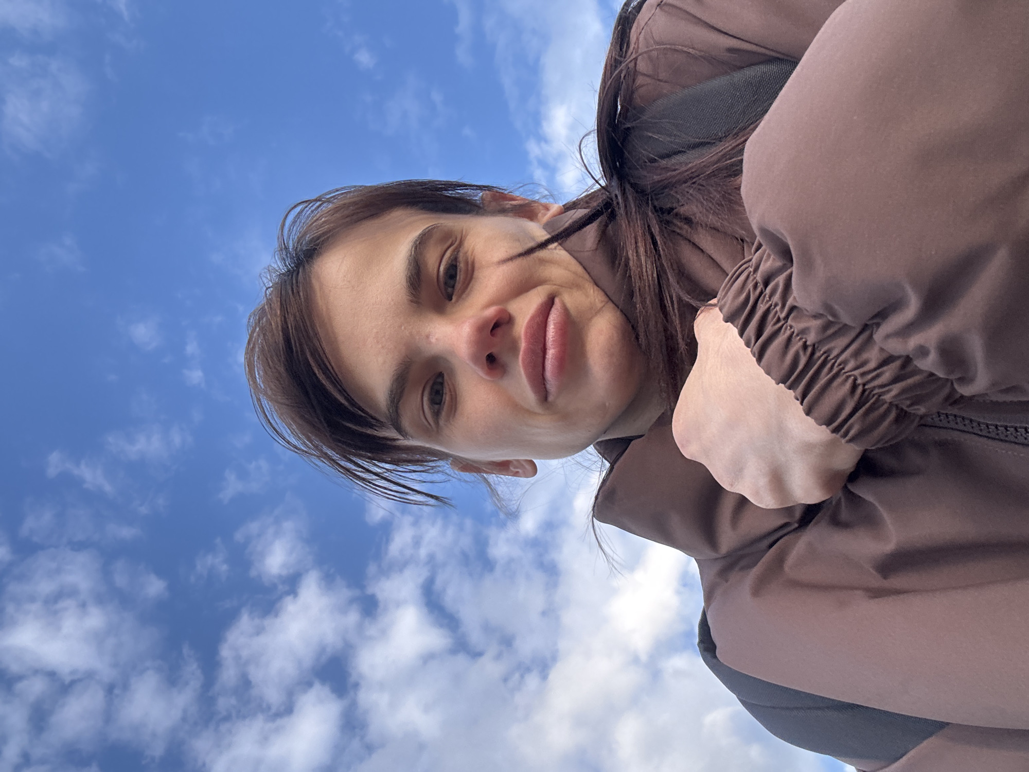 An outdoor selfie captures a young woman from a low angle, looking downwards at the camera. She has medium-length dark brown hair, parted on the side, with some strands falling across her forehead. Her expression is a subtle smirk or soft smile, with her lips slightly pursed and eyes open, looking towards the viewer. She is wearing a brown, seemingly puffy jacket with an elasticized cuff visible on her right arm. A dark strap, possibly from a backpack or bag, is visible behind her shoulder. The background is a bright blue sky with scattered white, fluffy clouds, indicating daylight and fair weather. The image is rotated, giving the impression that the person is positioned above the viewer. No specific location cues are visible within the image to confirm the provided context of Rostov-on-Don, Russia, nor is any text visible.