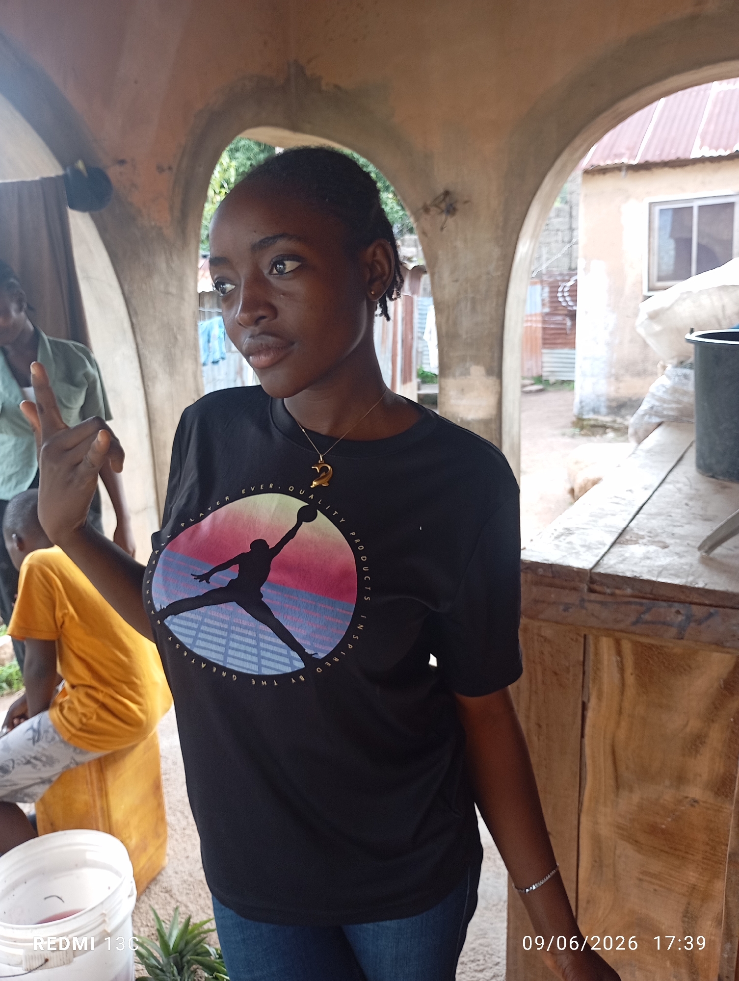 In Jos, Nigeria, a young woman stands under an arched entryway, her gaze directed to the left. She wears a black t-shirt with a circular graphic featuring a silhouette of a basketball player against a sunset, and a gold dolphin pendant necklace. To her left, a person in a green shirt is partially visible. Further to the left and slightly behind, another person is seated on a yellow container, wearing a bright orange shirt and patterned shorts. The environment suggests an outdoor setting, possibly a market or roadside stall, with a wooden counter and structure to the right. The background shows corrugated metal sheeting and a building with a red roof. A white bucket and some foliage are visible in the foreground. The date stamp "09/06/2026 17:39" is overlaid on the bottom right of the image, indicating a timestamp.