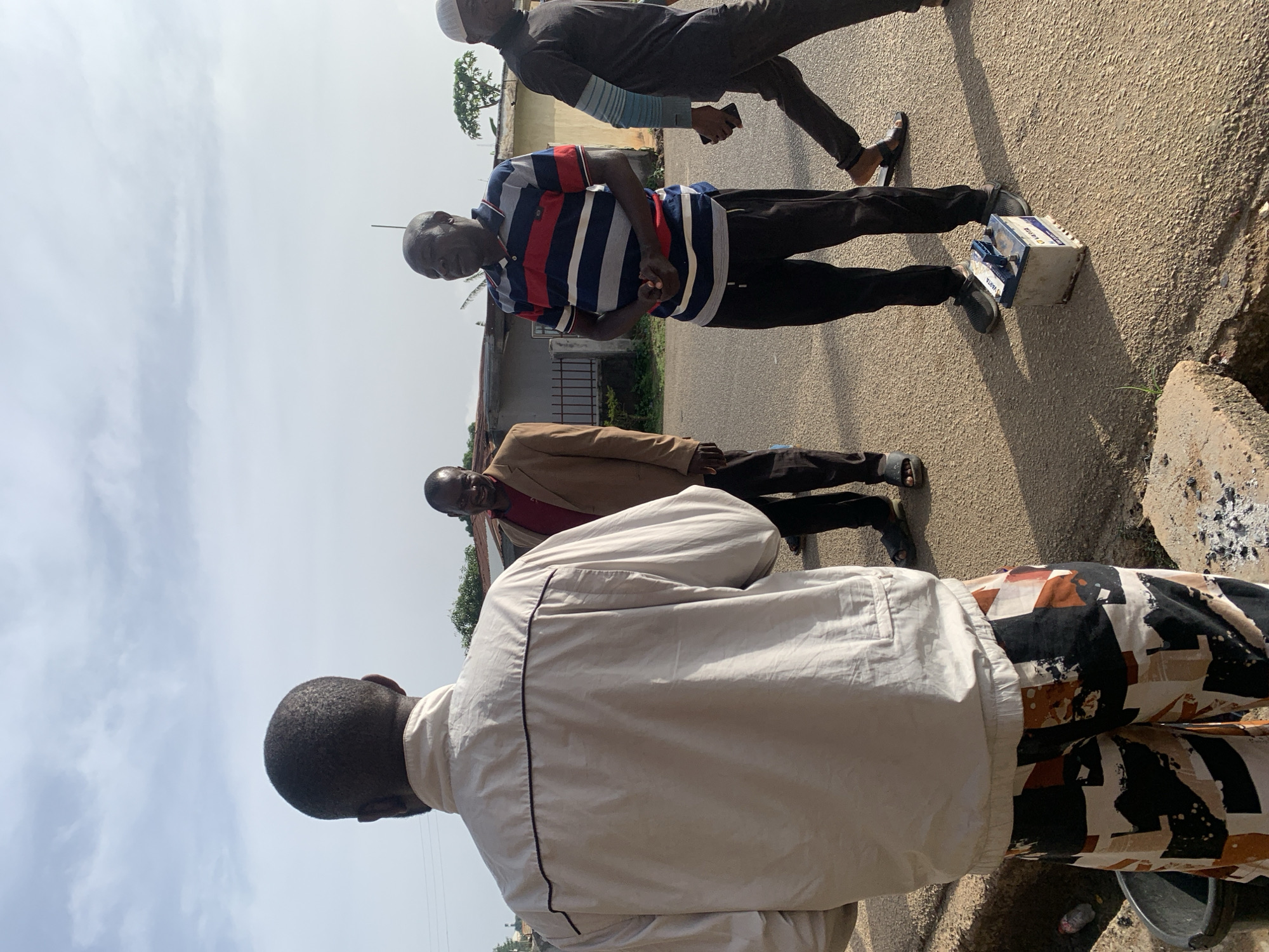 The image shows four men outdoors, seemingly on a street or road in Jos, Nigeria. In the foreground, from the back, a man wears a light-colored jacket over patterned pants. To his right, a man in a tan suit jacket and red shirt stands facing the camera, smiling. Next to him, a man in a striped polo shirt and black pants is facing forward with his hands clasped. To the far right, a man wearing a dark outfit and a white cap is walking away from the camera. A white and blue battery sits on the ground in front of the man in the striped shirt. The background consists of buildings and trees under a cloudy sky.