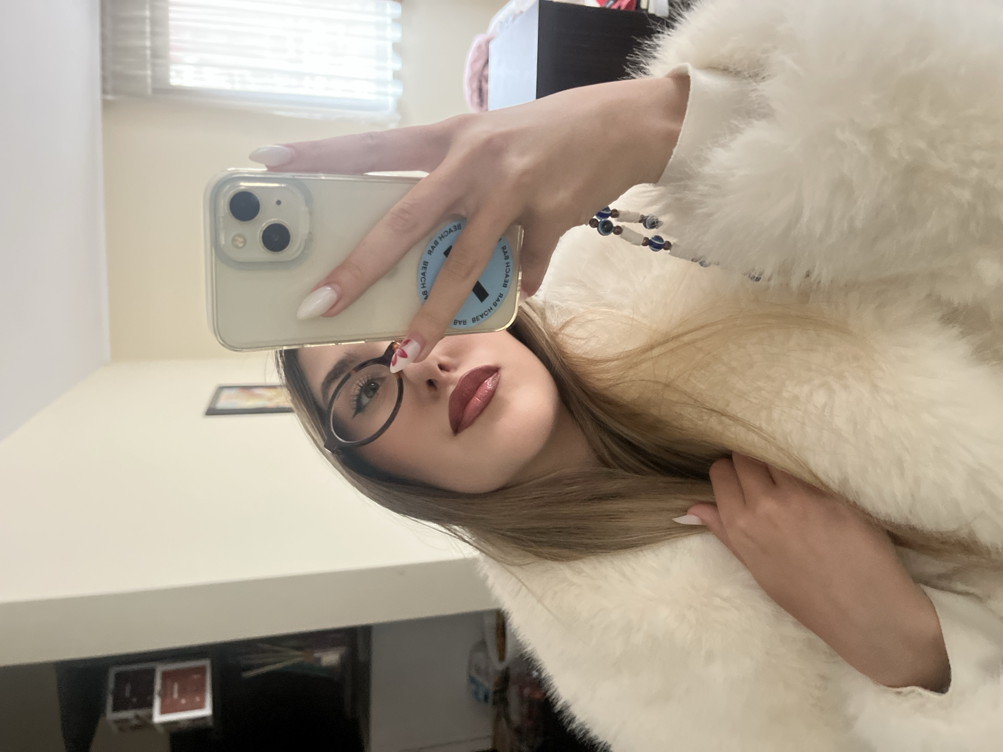 The media file shows a close-up, waist-up mirror selfie of a young woman, presented rotated 90 degrees clockwise from a typical upright orientation. She has long, light brown hair and is wearing a white, fluffy faux fur coat. Her face is turned slightly towards the viewer, featuring dark-framed glasses and reddish-brown lipstick.
Her right hand, adorned with long white almond-shaped nails, holds a light-colored smartphone (likely an iPhone) with a clear case, positioned to reflect her image. A blue and white beaded bracelet, possibly featuring evil eye beads, is visible on her right wrist. On the back of the phone, a blue PopSocket or phone grip is attached, displaying text that, when read as a mirror image, spells out "BEACH BAB".
The background, visible through the mirror reflection, includes an indoor room with light-colored walls, horizontal window blinds, and dark furniture. A framed picture is partially visible on the wall in the upper left. The lighting is bright and even, consistent with an indoor daytime setting in Sofia, Bulgaria. The woman has a neutral to slightly posed expression.