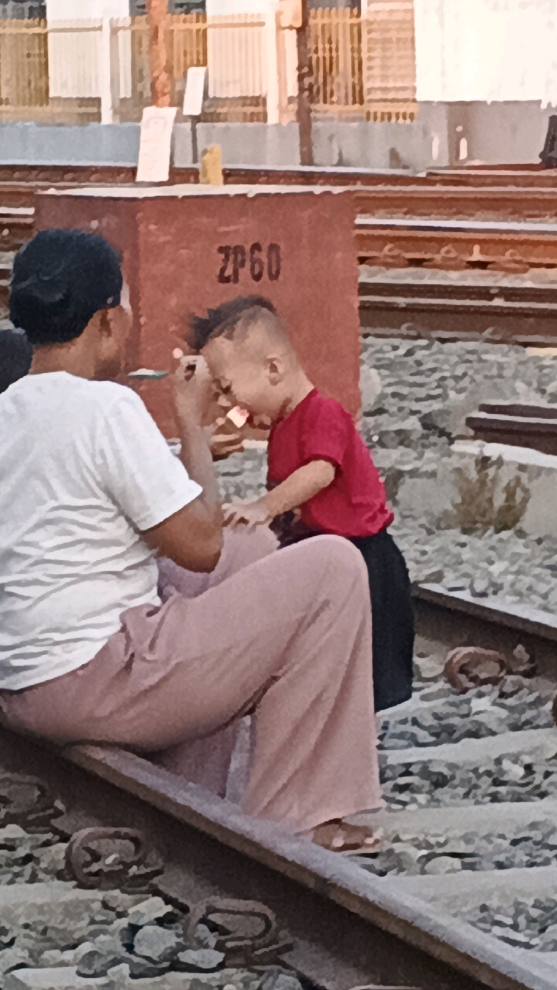 In Semarang, Indonesia, two individuals are present on a railway track. One is an adult, positioned to the left, wearing a white t-shirt and light pink pants. They are facing away from the camera, towards a child who is positioned in front of them and to the right. The child, dressed in a red shirt and black pants, is leaning forward, their mouth open as if eating or about to eat something held by the adult. The adult's left hand is near the child's face, holding what appears to be a piece of food. The child's right hand is outstretched. They are seated on a railway track, surrounded by gravel and ballast. In the background, a reddish-brown rectangular structure with the markings "ZP60" is visible, along with other railway tracks. Further in the background, buildings with a light-colored facade and orange fencing can be seen. The lighting suggests it is daytime, possibly late afternoon, given the warm tones in the image.