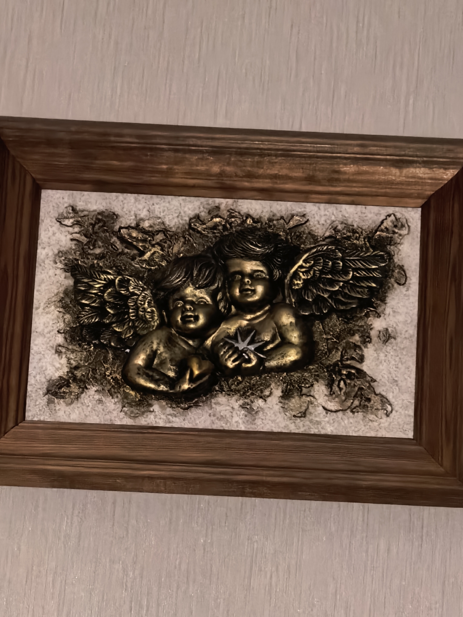 A framed relief sculpture of two cherubs is displayed on a textured, off-white wall. The sculpture is rectangular and appears to be made of a metallic material, possibly brass or bronze, with a golden sheen. The cherubs are depicted with wings spread, seated close together. One cherub is on the left, looking forward with its hands clasped. The cherub on the right is slightly turned towards the left cherub, holding a star-shaped object between its hands. The background of the sculpture is a textured, light-colored material, possibly plaster or stone, with darker, abstract elements suggesting foliage or clouds. The frame is made of wood with a dark, grainy texture, giving it a rustic appearance. The lighting in the image is subdued, casting subtle shadows that highlight the relief details of the sculpture. The overall scene suggests a decorative piece in an indoor setting, likely a home or a gallery.
