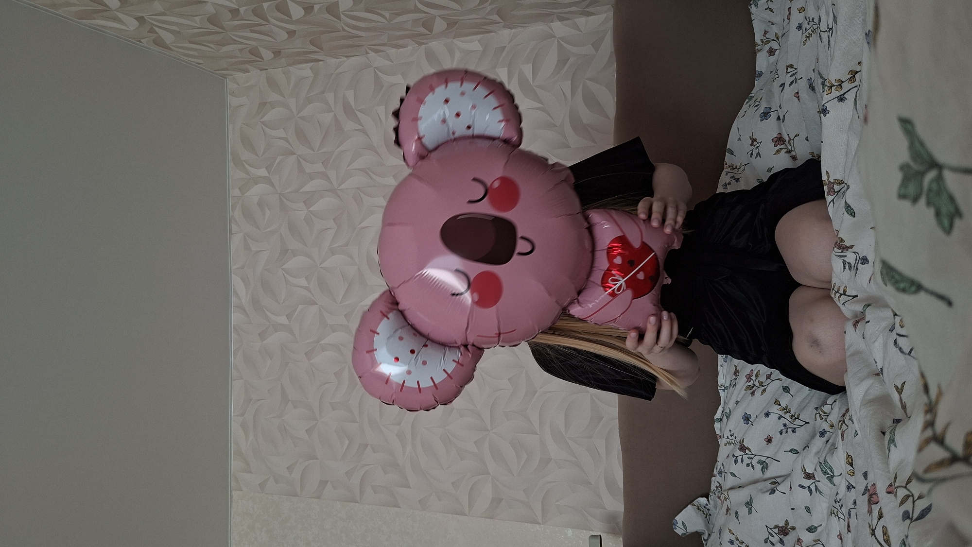 A person is sitting on a bed in a room, holding a pink koala-shaped balloon. The room has wallpaper with a geometric pattern on one wall and a plain wall on the other. The bedding has a floral print. The location is Chaikovsky, Russia.