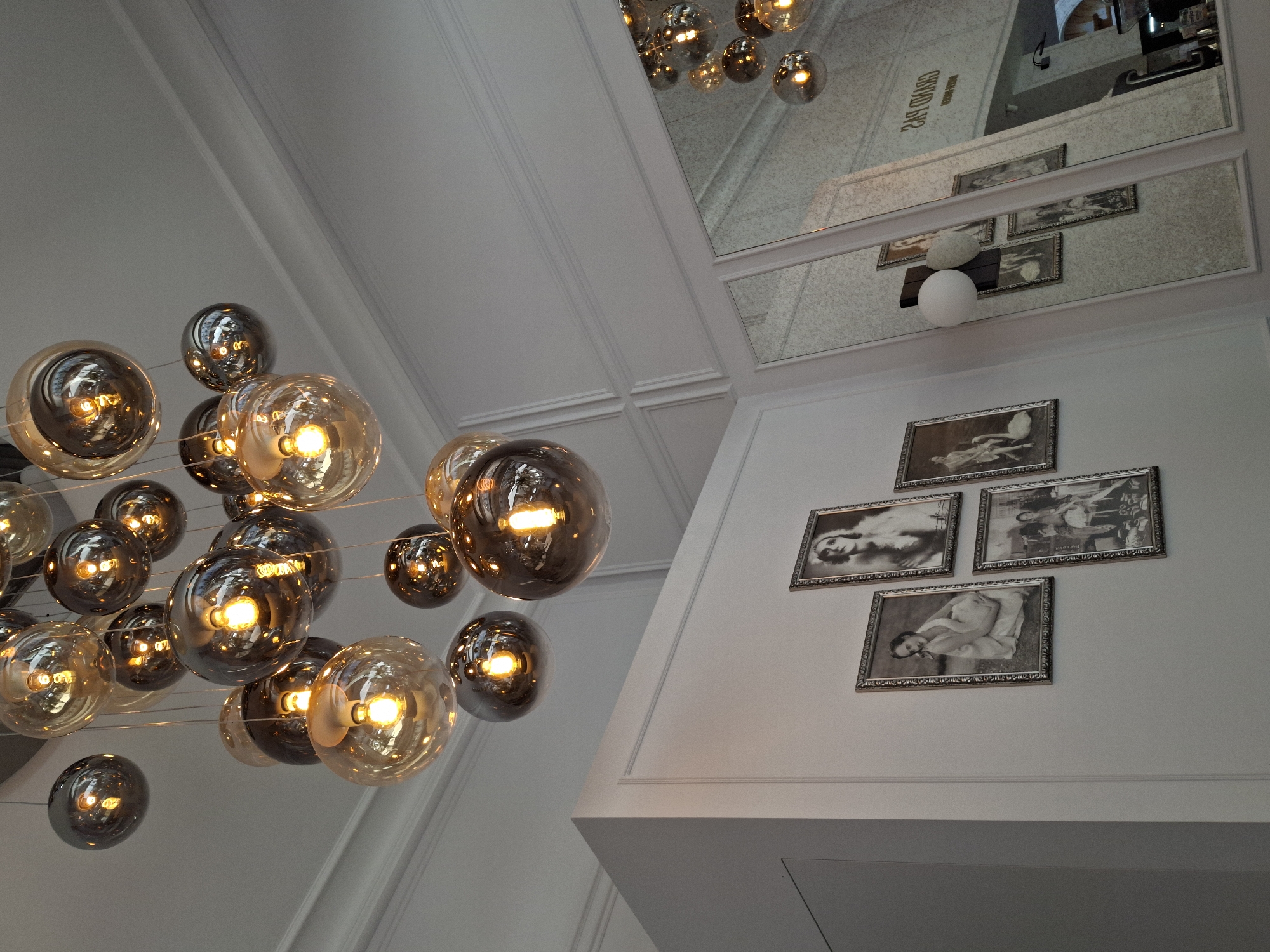 The image shows an interior scene with a focus on a modern chandelier made of multiple spherical glass bulbs containing lights. The chandelier hangs from a white, paneled ceiling. On the left wall, there are several framed black and white photographs of people, including at least one of a woman in a fur-trimmed garment and another of a woman posing with a swan. To the right, a mirrored wall reflects more of the chandelier and the environment, including what appears to be a sign with text that reads "GRAND PAS." The overall impression is of an elegant, potentially upscale interior space. Based on the visible signage, this scene is located at the Grand Palais in Baden-Baden, Germany.
