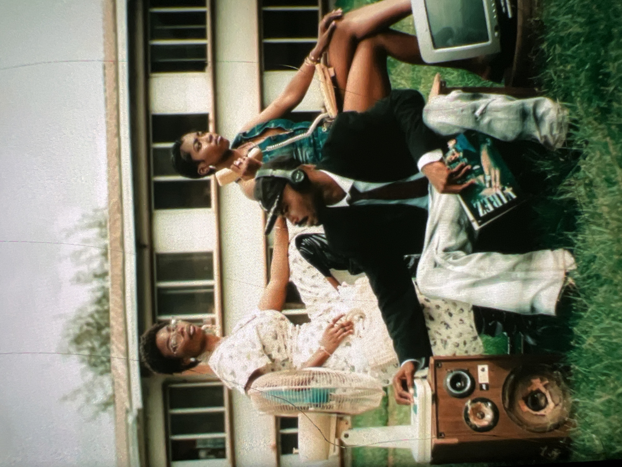 In Lagos, Nigeria, three individuals are posed outdoors in front of a two-story building with a row of dark-paned windows. The person on the far left, a woman wearing glasses and a floral dress, sits with her back to the viewer. Beside her, a man in a black suit jacket, white shirt, and maroon tie is seated, wearing headphones and a baseball cap. He holds a magazine titled "VIBEZ" on his lap. To the right, another woman in a green low-cut top and gold jewelry sits, holding a beige rotary telephone to her ear. A white electric fan is positioned to the left of the man, connected to a wooden speaker box. To the right of the woman with the phone, a small vintage television sits on a wooden stool. The scene is set on a grassy lawn under a cloudy sky.