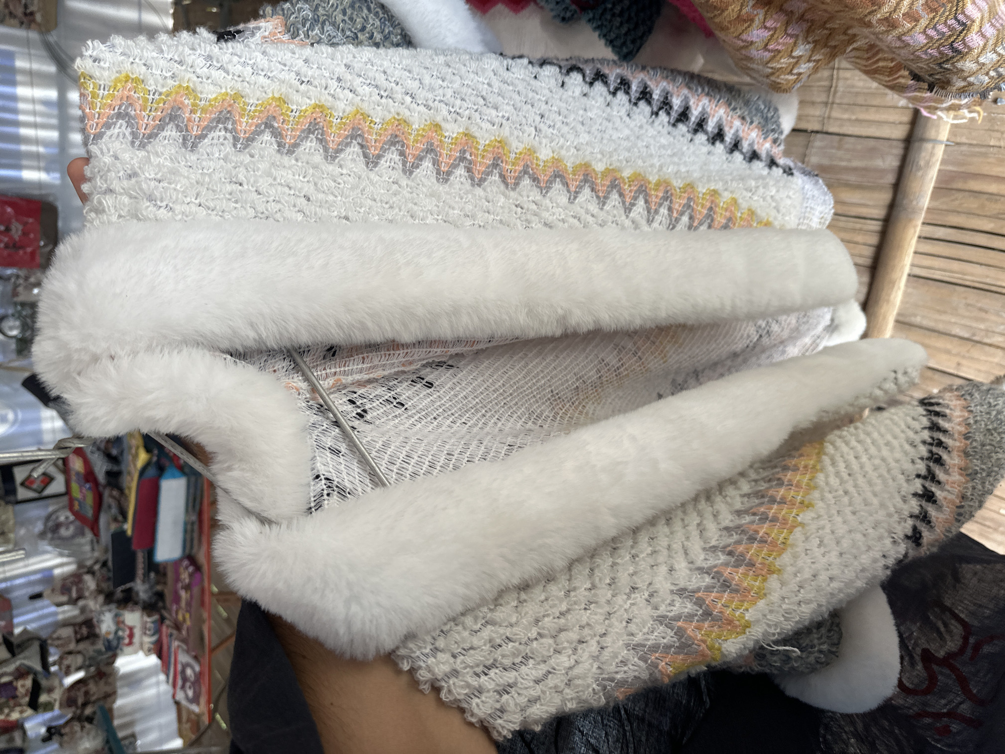 This is a close-up, eye-level shot of a person's arm and torso holding a fluffy white-trimmed sweater with a zig-zag pattern of pink, yellow, and gray. The sweater is hanging on a metal rack in what appears to be an outdoor market stall in Mai Chau, Vietnam. In the blurred background, other textiles, bags, and market goods are visible, suggesting a vibrant commercial setting. The wooden plank floor and the natural light indicate an authentic, possibly rural, market environment.