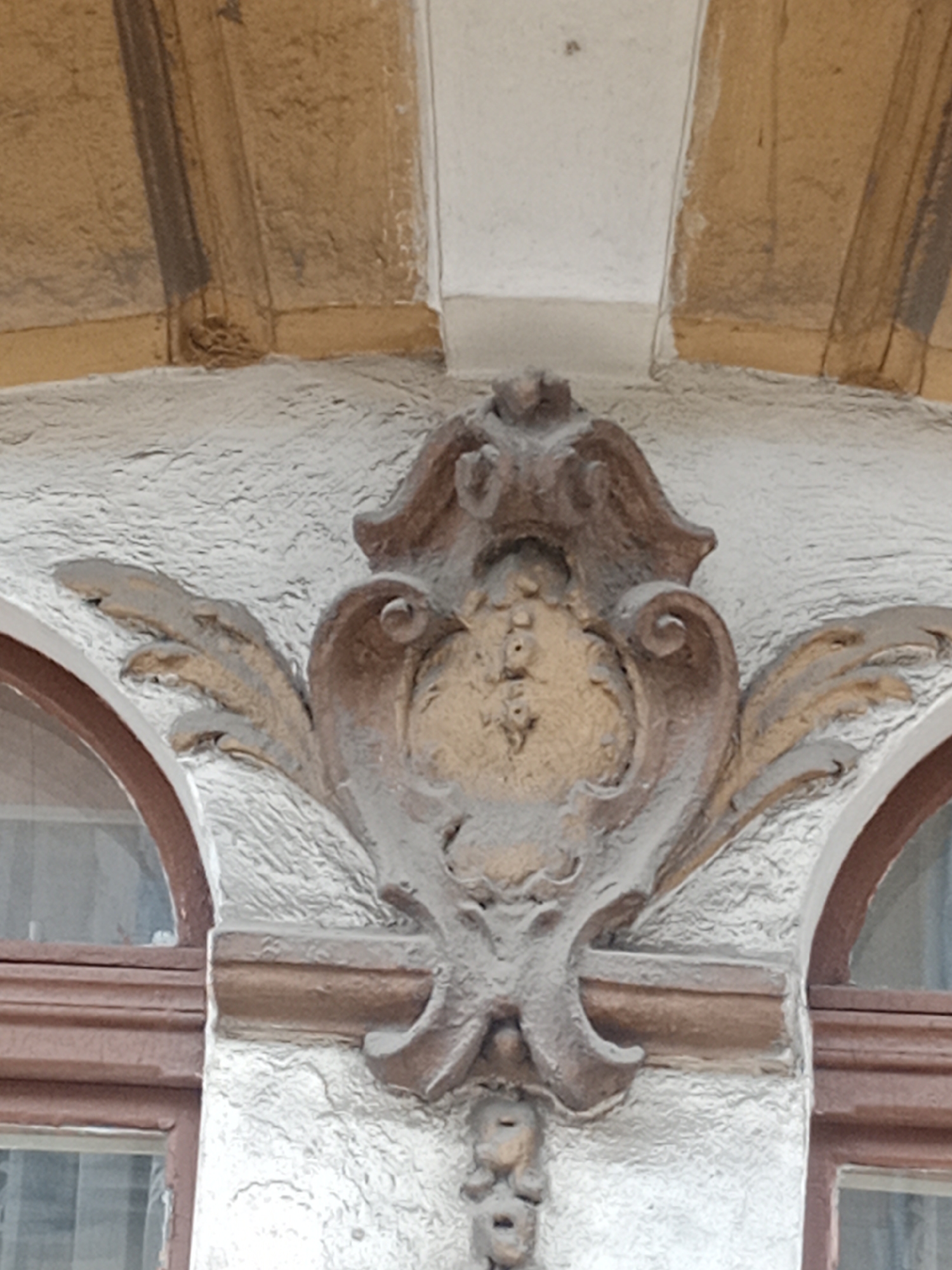 The image shows a close-up, eye-level exterior shot of ornate architectural decoration above a window on a building. The decoration is a symmetrical, carved element with a central shield-like shape featuring a pattern of small, circular indentations. Flanking the central element are stylized, wing-like forms and volutes, all rendered in a weathered, brown-toned material. The background consists of textured light-colored stucco and the underside of an arched architectural feature, also in a light brown hue. A portion of a brown window frame and the arched glass of a window are visible to the left and right. This architectural detail is located on the building at Plac Wolności 1 in Bydgoszcz, Poland.