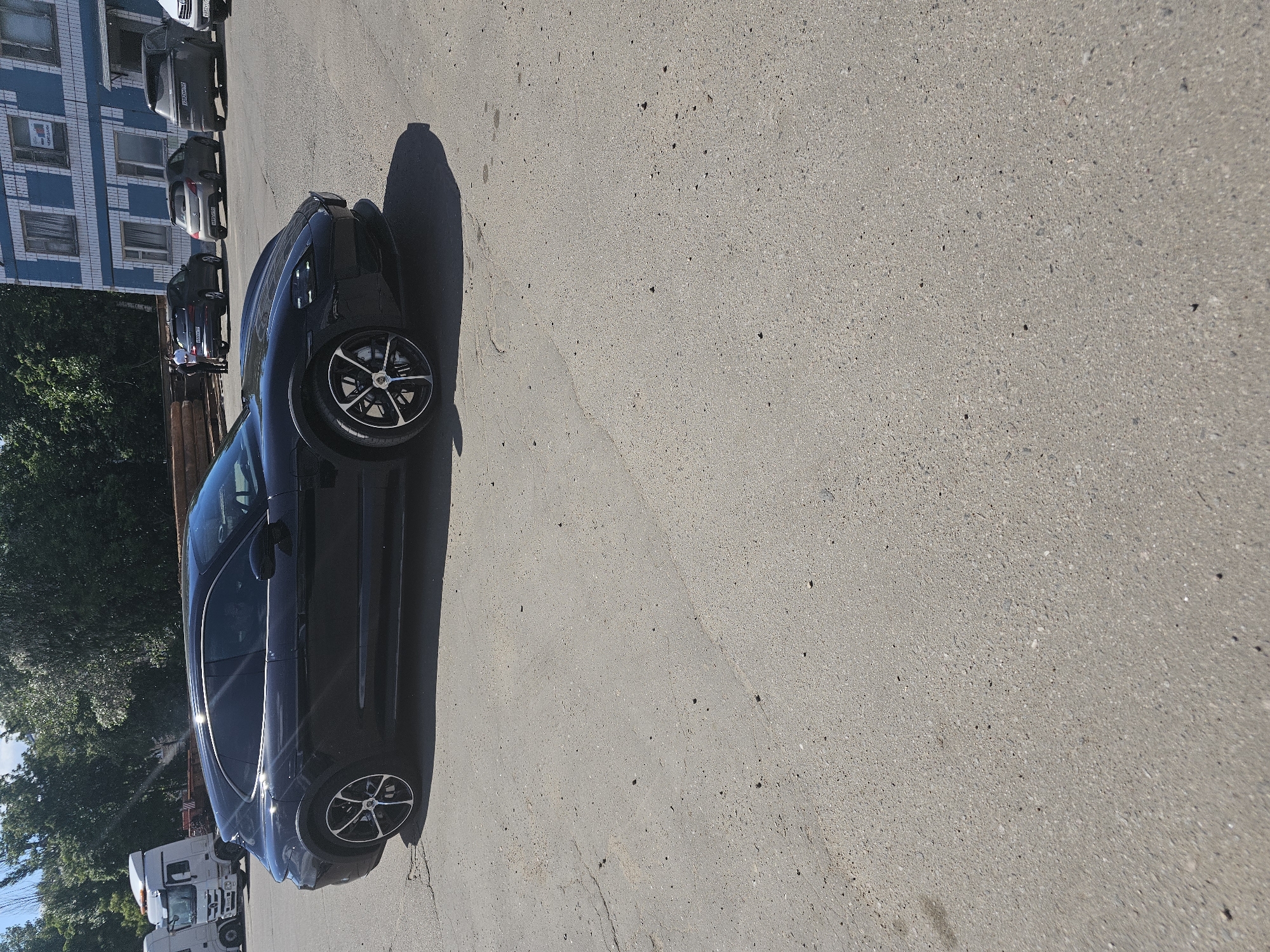 The image, oriented horizontally, features a dark-colored (likely black or dark blue) car, resembling a Porsche Taycan, parked on a light gray asphalt or concrete surface. The car is positioned prominently in the foreground on the right, angled slightly towards the left. It displays distinctive black and silver multi-spoke wheels with a crest visible in the center caps, characteristic of Porsche.
Bright sunlight casts long shadows from the car, indicating daytime, possibly morning or late afternoon, with clear weather. In the background, on the left side of the frame, several other cars are parked in a line, mostly dark-colored sedans or hatchbacks. One of these cars, a gray vehicle, has a license plate with Cyrillic characters, consistent with the Moscow, Russia context. Further back, a light blue building with a patterned facade and multiple windows with white frames stands. A white sign with illegible text is visible on one of the building's windows. Green trees and foliage are also present on the far left, along with a partially visible white semi-truck.
The ground is a textured, light gray surface. No people or specific activities are visible; the scene depicts parked vehicles in what appears to be an urban or suburban setting.