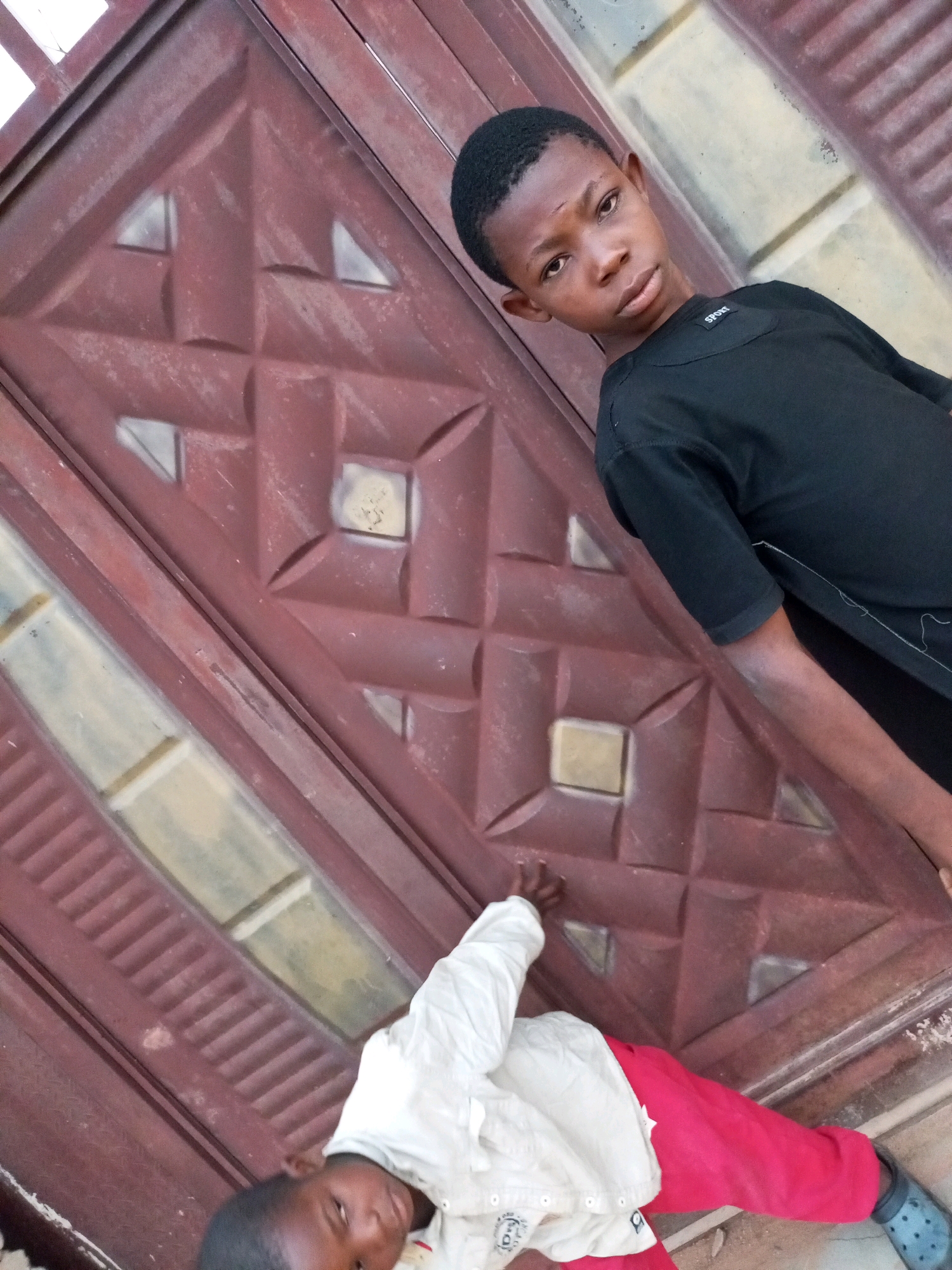 Two young boys are positioned in front of a large, dark red metal door with a geometric pattern. The boy on the right, wearing a black t-shirt, is standing and looking towards the camera with a neutral expression. The boy on the left, wearing a white shirt and red pants, is lying on the ground, facing away from the camera. The scene appears to be outdoors, with a textured surface beneath the boys. The location is identified as Tempure, Nigeria.