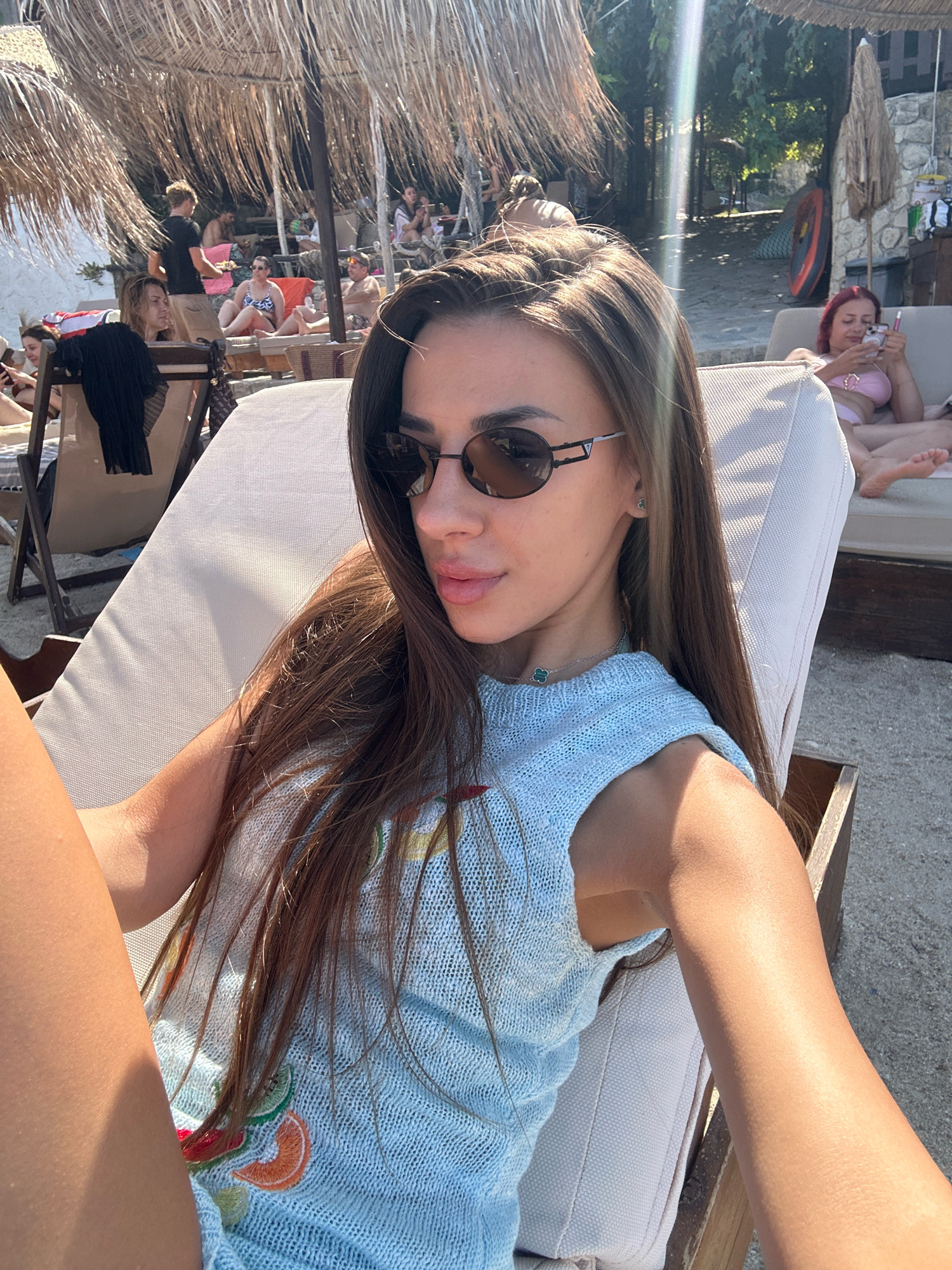 A young woman with long, dark brown hair and sunglasses is relaxing on a beach chair at a beach in Kassandra Municipal Unit, Greece. She is wearing a light blue, sleeveless knitted top with embroidered fruit designs. The background shows other beachgoers, umbrellas made of straw, and lounge chairs. The sun is shining, creating shadows and highlights on the scene.