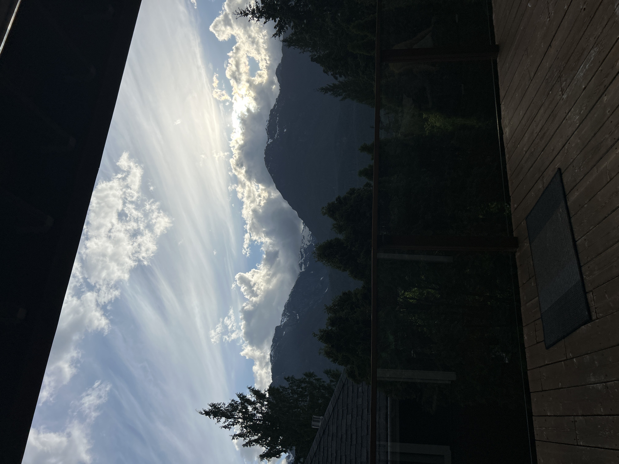 The photograph captures a scene in Squamish, Canada, showcasing a view of mountains framed by a deck or balcony setting. The sky is filled with white, streaky clouds against a blue backdrop, suggesting a partly cloudy day. A large mountain range dominates the background, partially obscured by the clouds. In the midground, trees add to the natural scenery. The foreground includes a wooden deck area, with a black mat or rug visible. To the left, a part of a building structure is present, possibly an overhang or roof. The image suggests a moment of observing the landscape from a residential or lodging setting in Squamish.