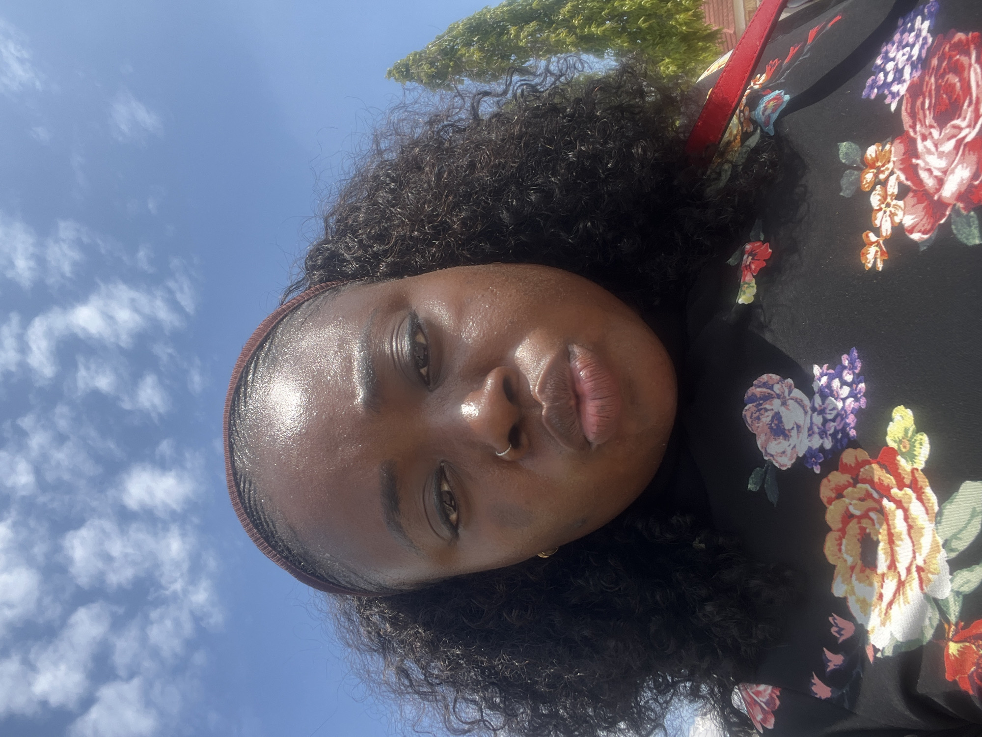 A dark-skinned female individual is depicted in an outdoor, upward-angled selfie, presumably in Abuja, Nigeria. She has dark, curly hair secured by a reddish-brown headband. Her skin exhibits highlights consistent with sun exposure or perspiration on the forehead, nose, and cheeks. She wears a small silver hoop nose ring on her left nostril and a gold hoop earring on her visible left ear. Her eyes are open, directed towards the camera, and her lips are pursed into a distinct pouting expression. She is dressed in a black top featuring large, multi-colored floral patterns in shades of red, orange, yellow, purple, and blue. A red strap is partially visible over her right shoulder, suggesting a bag or garment accessory. The background consists of a clear blue sky with dispersed white cumulus clouds. A tall, narrow green tree is visible on the right side, with a light-colored building partially discernible below it.