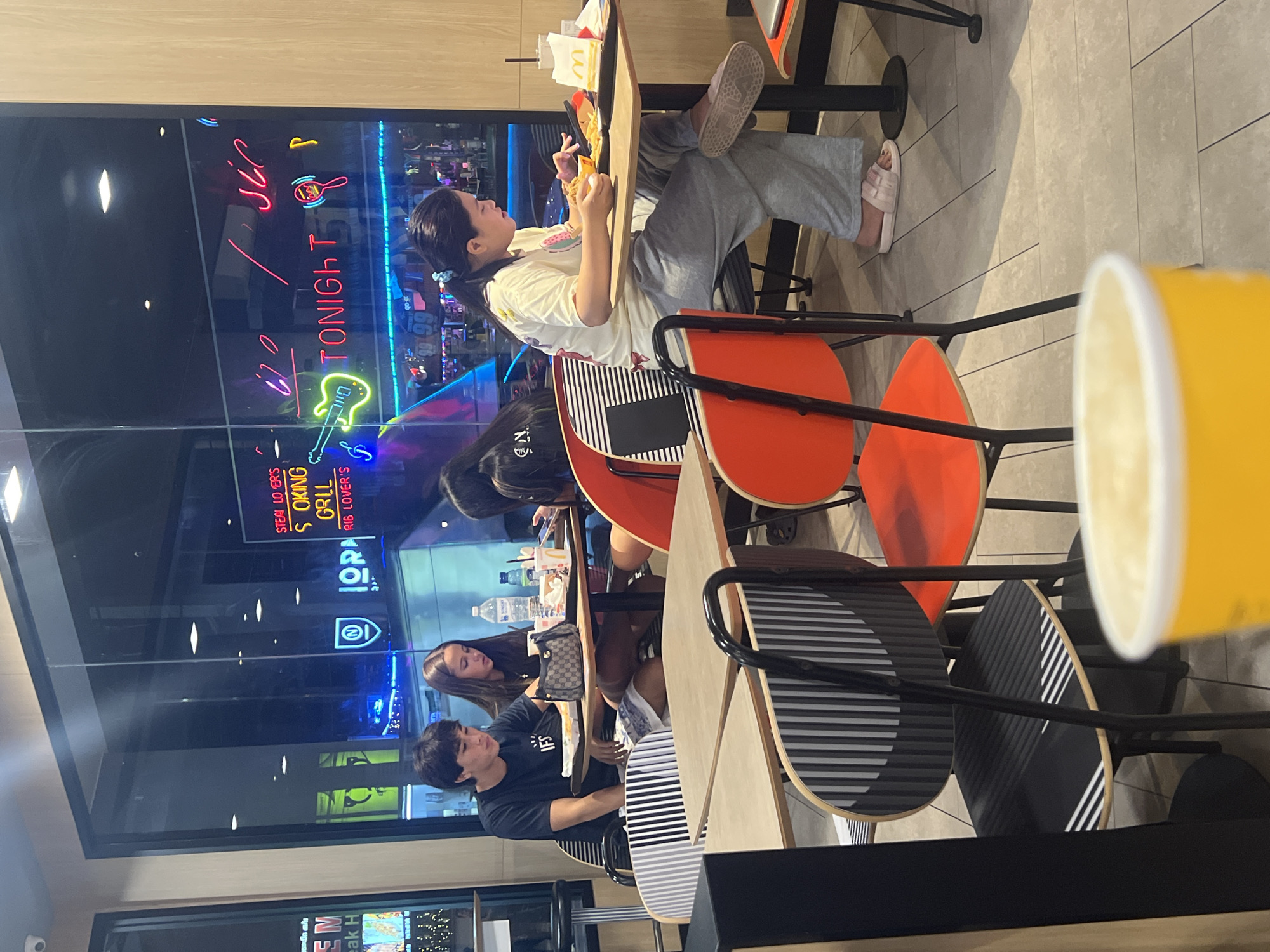 The image shows a group of people dining at tables inside a fast-food restaurant, possibly McDonald's, as evidenced by the iconic golden arches visible on a sign in the background. The scene is illuminated by artificial lighting, suggesting it could be evening or a dimly lit interior.
Several individuals are visible. In the foreground, a young person with dark hair, wearing a black t-shirt, is seated at a table, looking down. Beside them, a young woman with long brown hair, wearing a dark top, also appears to be eating or looking at something on the table. Further back, a young girl in a light-colored t-shirt and gray pants is sitting at a table with her legs propped up, holding a piece of food and looking at her phone. A second young woman in a dark t-shirt is seated at another table, with her head bowed.
The tables have distinctive red and black patterned seats and light-colored tabletops. Outside the window, neon signs are visible, including "TONIGHT" and a stylized electric guitar, hinting at a possibly lively or entertainment-oriented atmosphere outside the restaurant. A bright yellow cup is prominently placed in the foreground, partially obscuring the view of the other diners. The overall impression is of a casual dining experience.