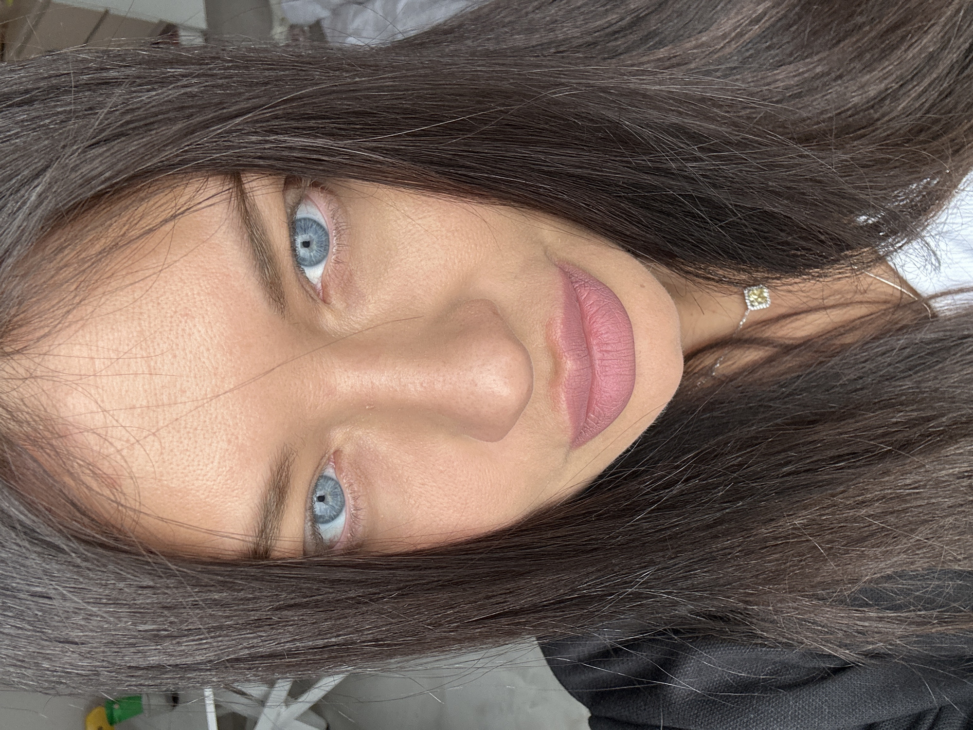 A close-up, high-detail portrait features a person's face, oriented on its side, focusing primarily on their left eye, nose, mouth, and surrounding hair. The subject has striking light blue eyes with visible pupils and irises. Their long, dark brown hair frames the face, with individual strands, some appearing lighter or gray, visible across the forehead and around the sides. The skin has a natural texture. The person wears subtle eye makeup and has light pink matte lipstick on their lips. A thin silver necklace with a small, square, yellowish-clear pendant is visible around the neck, partially obscured by hair on the right side of the frame. The background is indistinct and out of focus, suggesting an indoor setting. The expression is neutral, with a direct gaze towards the camera. No visible text is present in the image. This scene is set in Khimki, Russia.