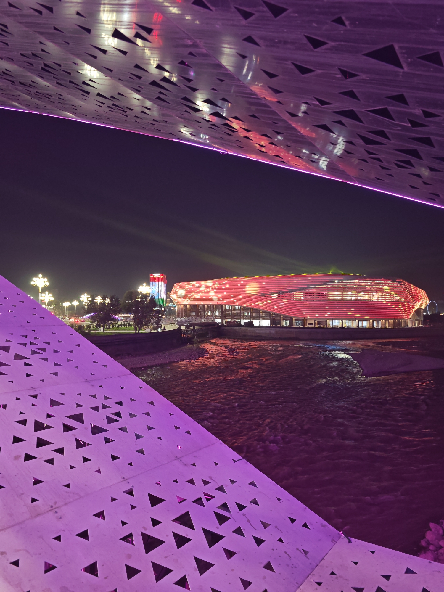 The image depicts an urban nighttime scene in Dushanbe, Tajikistan, likely near the Dushanbe Plaza or a similar modern architectural complex. In the foreground, a large, perforated structure with triangular cutouts is illuminated in a vibrant purple hue, forming a canopy or bridge element. This structure partially obstructs the view of the background.
Beyond the foreground, a river flows, its surface reflecting the colorful lights of the city and creating an undulating texture. Across the river, a modern stadium or large building is prominently featured, bathed in a dynamic red and pink light display. Horizontal lines of light emphasize its architectural form. To the left of the stadium, a tall building stands, displaying the colors of the Tajik flag (red, white, and green) through its illumination. Streetlights with ornate lamp heads line a pathway or road in the mid-ground, casting a warm glow. Trees are also visible in this area, some with subtle uplighting. The sky is dark, typical of nighttime, with faint light beams suggesting stage lighting or spotlights. The overall atmosphere is one of modernity and urban vibrancy, highlighted by the extensive use of colorful lighting. There are no people visible in the image.