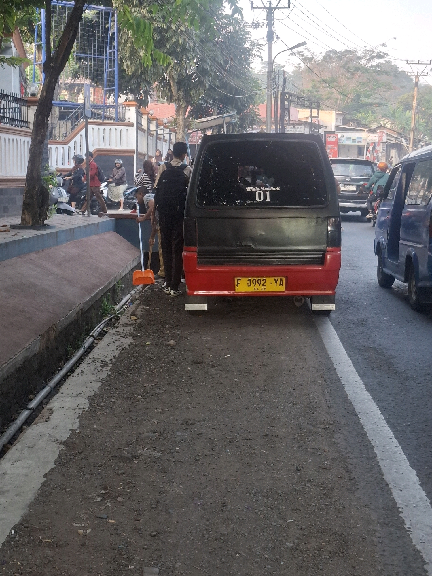 In Sirnagalih, Indonesia, an individual is sweeping debris from a roadside drain with an orange dustpan and a broom. The action takes place on a paved road, adjacent to a sidewalk with a blue border. A black and red van with a license plate F 1992 YA is parked on the right side of the road. To the left of the van, several people are seen standing and walking on the sidewalk, some of whom are on motorcycles. In the background, buildings and trees line the street, and utility poles with numerous wires are visible. The overall scene suggests a typical morning in a residential or commercial area.