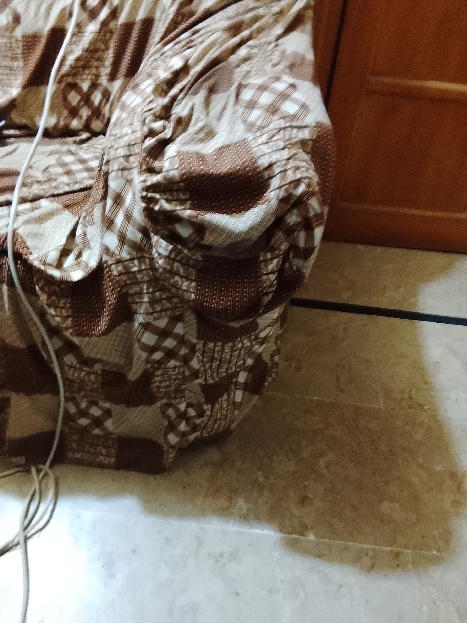 The image depicts a section of a patterned brown and beige upholstered sofa or armchair, positioned next to a wooden door frame. A white cord is visible to the left of the sofa. The scene is indoors, with a light-colored, possibly tiled floor. The environment is not distinct enough to determine the specific location within Multan, Pakistan.