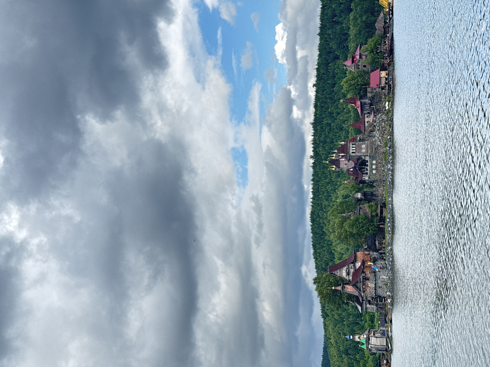 The image depicts a lakeside view in Satka, Russia, on a cloudy day, likely in the late afternoon. The scene features a row of rustic, fantasy-themed buildings nestled along the shoreline, backed by a dense, dark green forest. These structures have distinctive red-tiled roofs and appear to be made of stone and wood, resembling a medieval or fairytale village. Several buildings have turret-like elements, adding to the whimsical aesthetic.
The water in the foreground is rippling, suggesting a gentle breeze. The sky is a mix of dark, heavy clouds and patches of bright blue sky, indicating dynamic weather. One of the buildings has a bright yellow and blue object on its roof, possibly an umbrella or decoration. A small, dark bird is visible soaring in the distance against the cloudy sky.
The overall atmosphere is one of natural beauty blended with a touch of fantasy, creating a unique and intriguing landscape. No text is visible in the image.