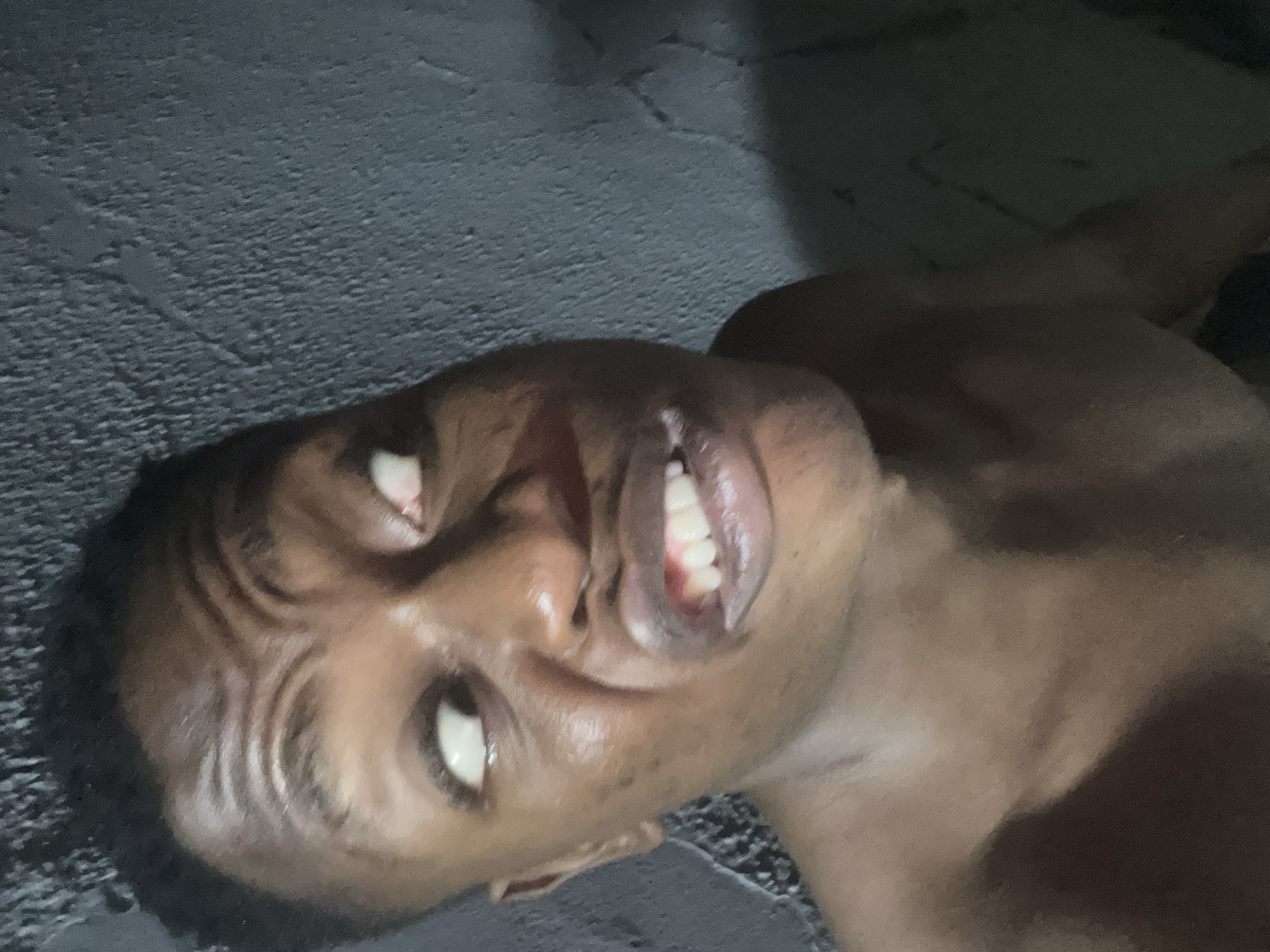 A close-up, portrait-oriented image features an individual, likely male, with dark skin, captured in an exaggerated facial expression. The scene is set in Ikorodu, Nigeria.
The subject's eyes are rolled upwards and to the right, showing predominantly white sclera; a reddish tint is visible in the corner of the left eye. The forehead is deeply furrowed with multiple prominent horizontal wrinkles. The mouth is open wide, baring both upper and lower white teeth and reddish gums. The lips are full and dark. A small blemish or mark is visible on the left cheek. The individual is bare-chested, with the upper torso and shoulders visible; the skin appears slightly glossy, possibly from moisture or natural oils.
The background primarily consists of a dark, textured wall, consistent with rough plaster or concrete, appearing dark grey or charcoal in color. This wall surface is particularly evident on the right side of the frame and recedes into deeper shadow towards the lower right. The left side of the frame is also dark, suggesting a similar wall or an unlit area. Lighting is direct and uneven, highlighting the forehead, nose bridge, lips, and shoulders, while casting pronounced shadows in other areas, especially on the left side of the face and the lower right background.
